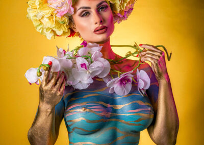 A breathtaking award-winning fine art body paint portrait by Victoria Machin Photography, featuring a model adorned with a vibrant floral headpiece, metallic body art, and ethereal tulle. The luxury fine art photography composition blends fashion, beauty, and artistic expression, making it a visionary Vogue-inspired masterpiece.