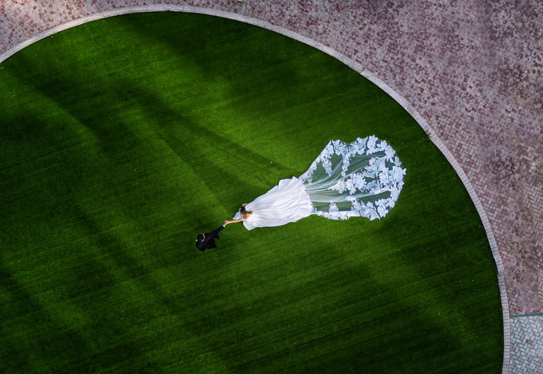 Drone Wedding Portrait at Trump Towers Sunny Isles | Victoria Machin Photography Aerial view of a bride and groom holding hands on a green lawn, with the bride's floral lace veil flowing behind, captured by drone at Trump Towers Sunny Isles.