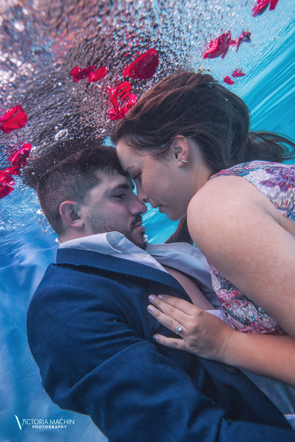 Engaged couple floating underwater with red rose petals, gazing into each other’s eyes during a romantic underwater photoshoot in Miami by Victoria Machin Photography.