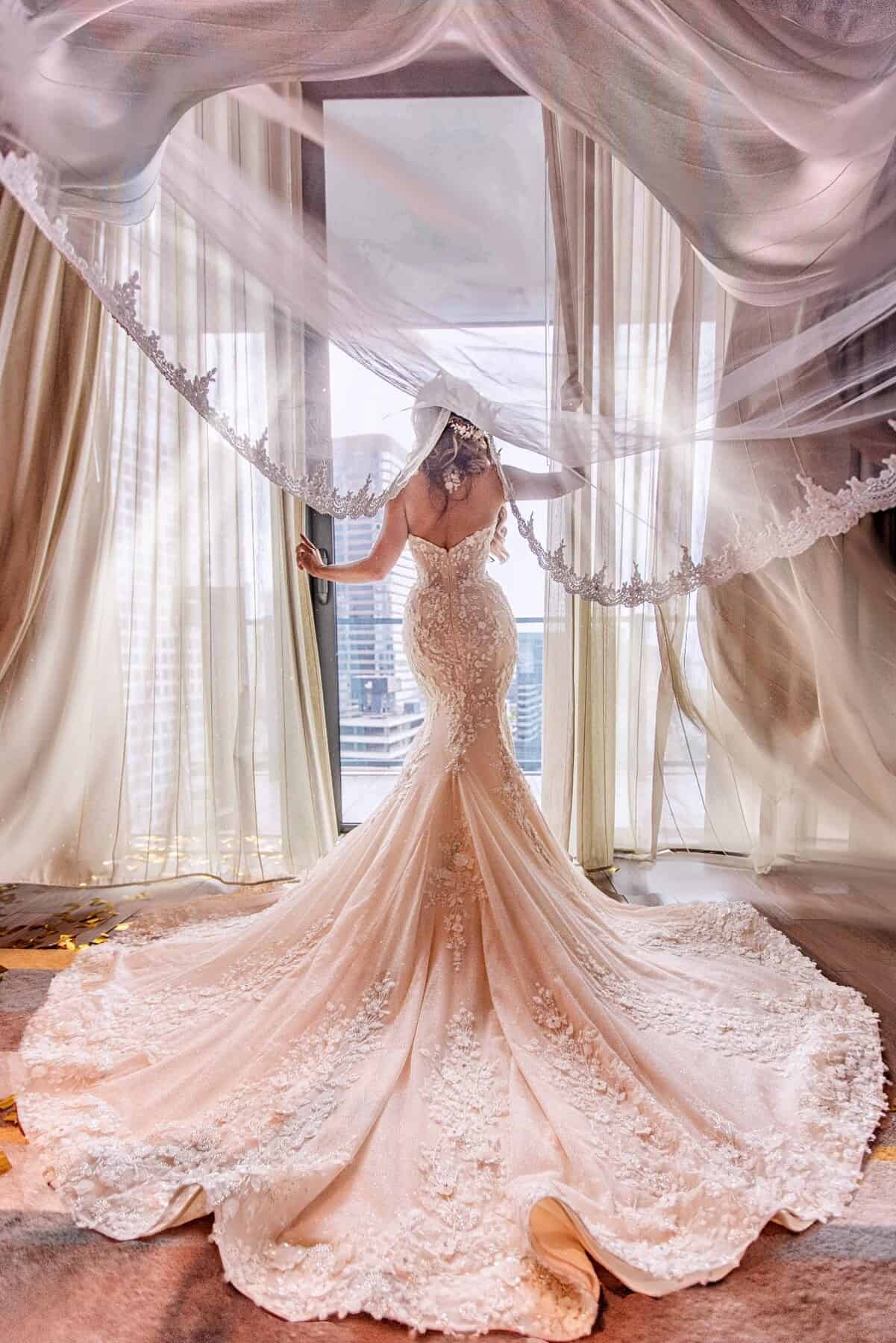 Bride from behind as her lace-edged cathedral veil billows above her, gown train spread across the floor before a city-view window