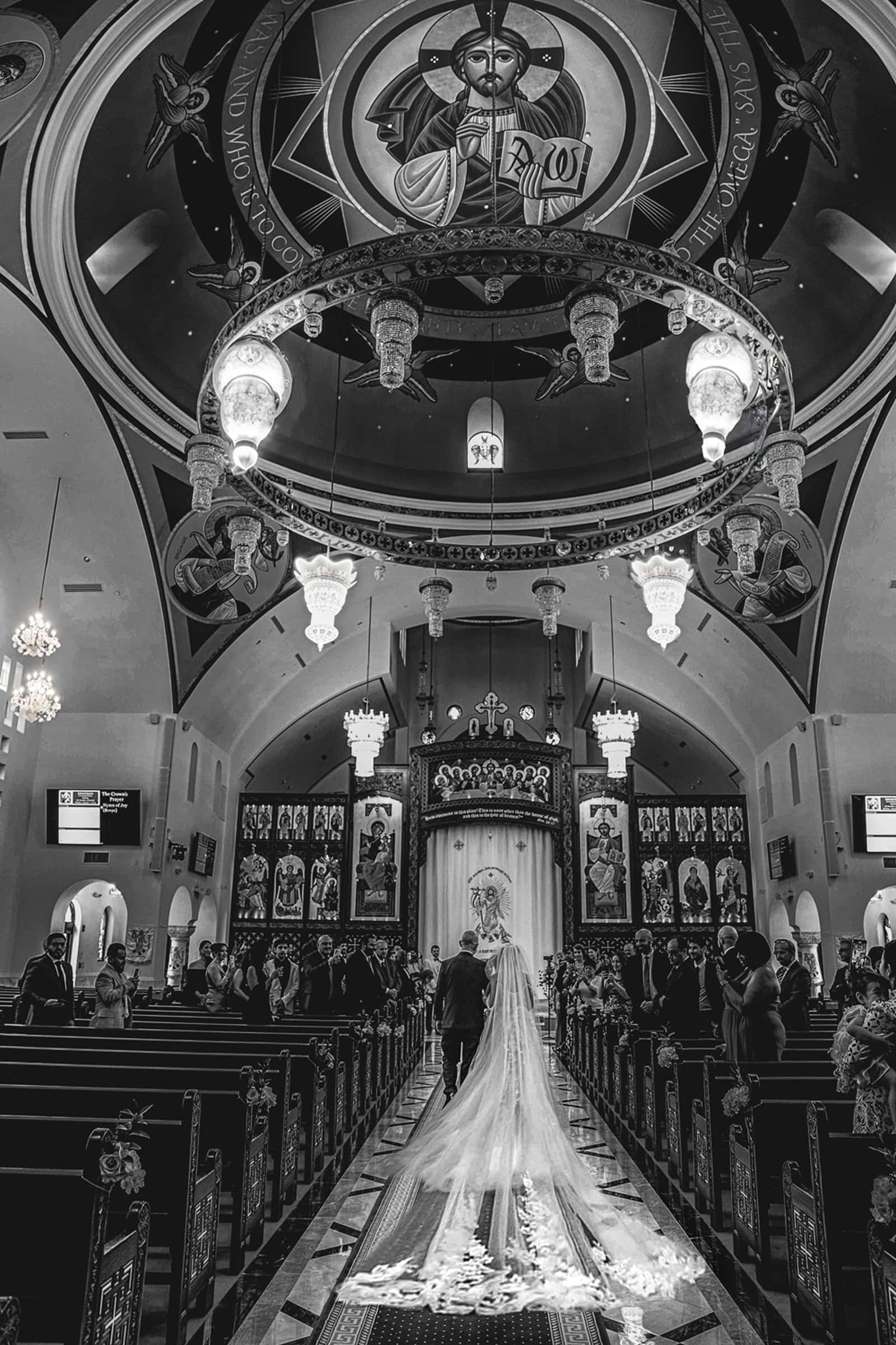 Bride and her father walking down the aisle of a domed church beneath a grand chandelier, in black and white