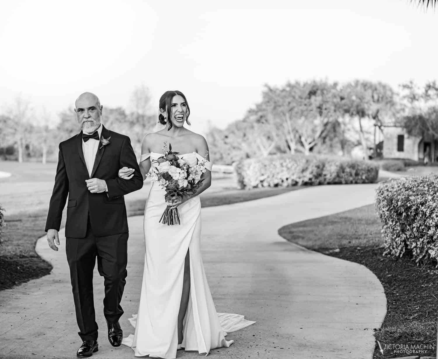 Bride laughing with joy as she walks arm in arm with her father, in black and white