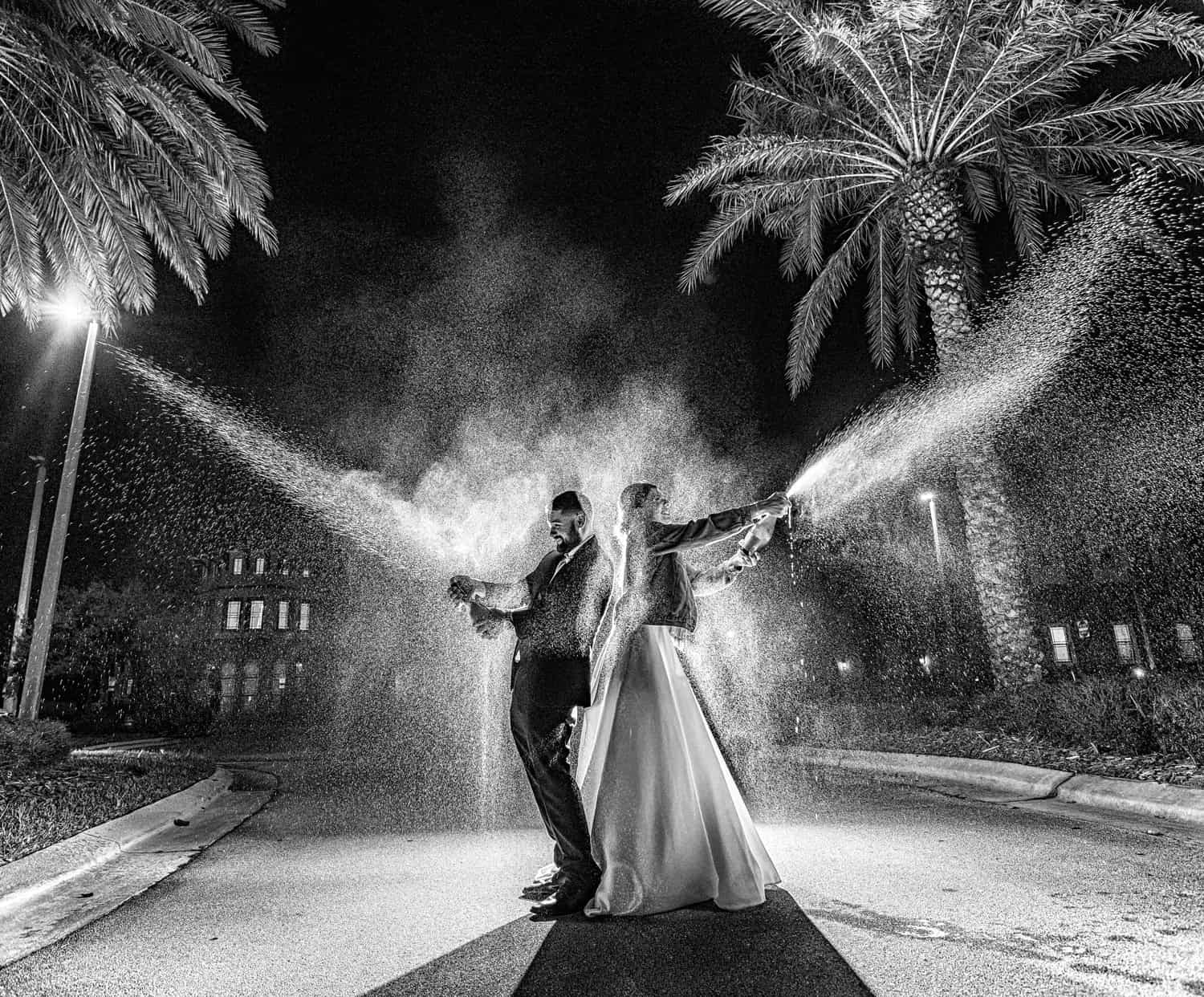 Bride and groom spraying champagne beneath palm trees at night, black and white