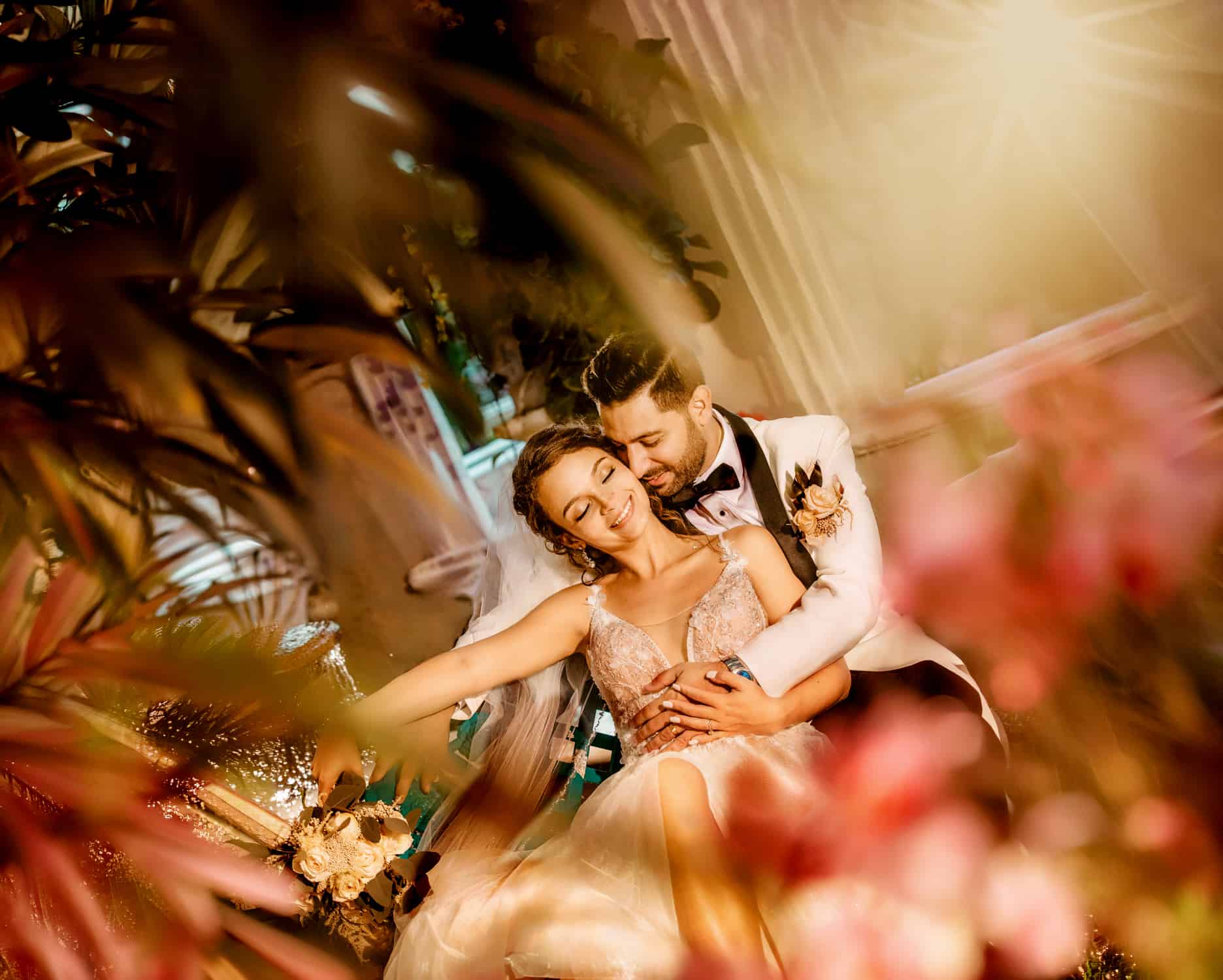 Groom embracing his smiling bride from behind, photographed through soft blush florals in warm light