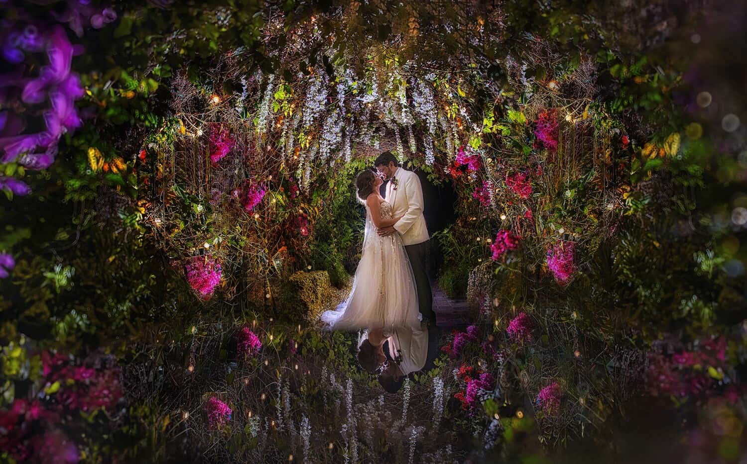 Bride and groom embracing beneath a floral archway lit at night, reflected in water below