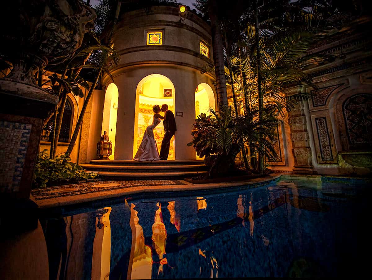 Bride and groom kissing beneath lantern-lit arches, reflected in the pool at night