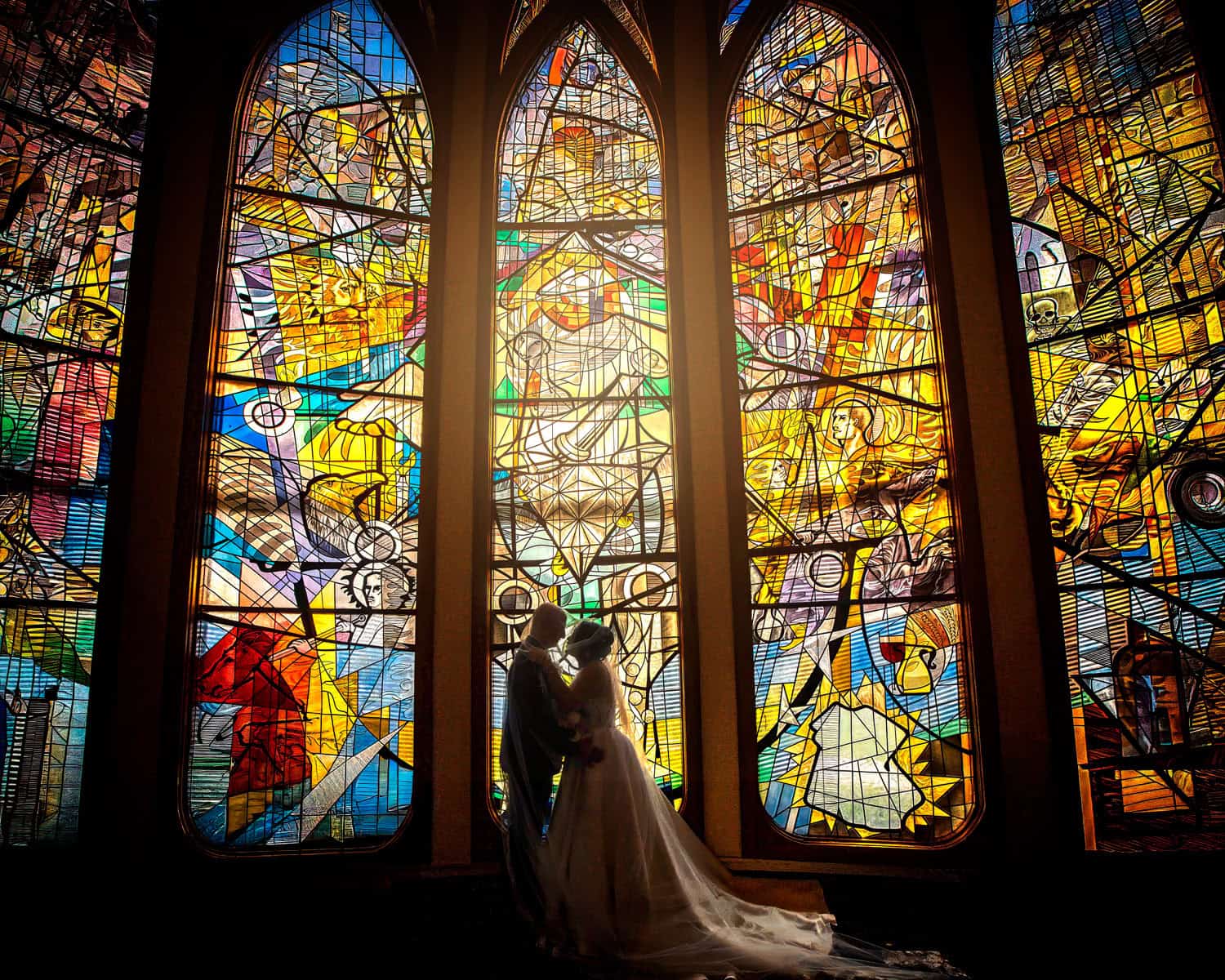 Bride and groom silhouetted in a kiss before towering stained-glass windows