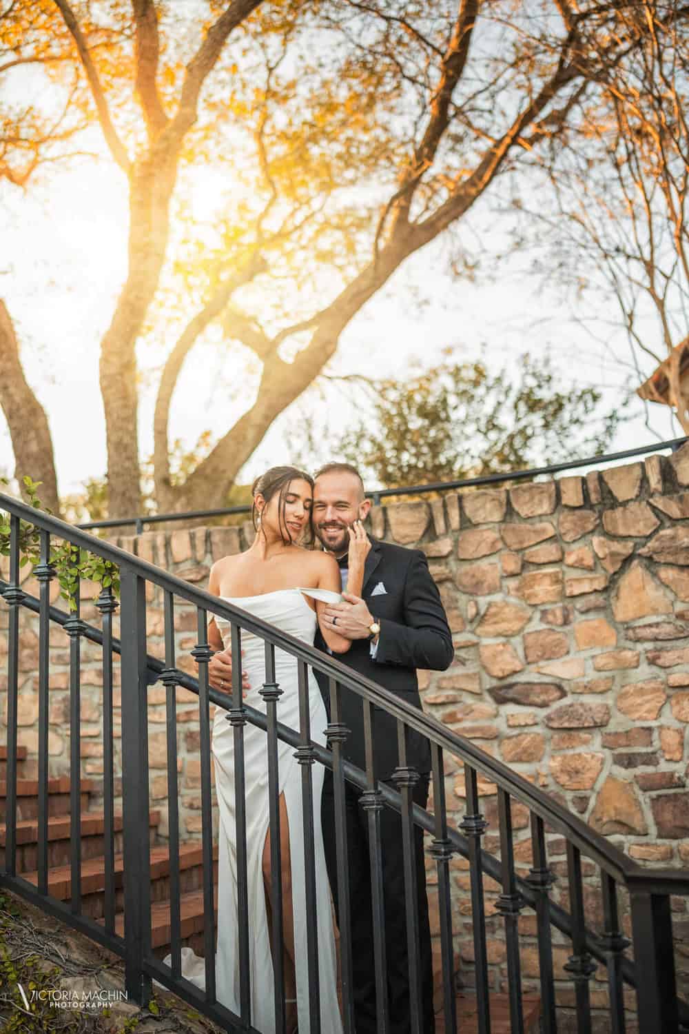 Bride and groom on a wrought-iron staircase against a stone wall, backlit by sunset through the trees, Parkland Golf Club