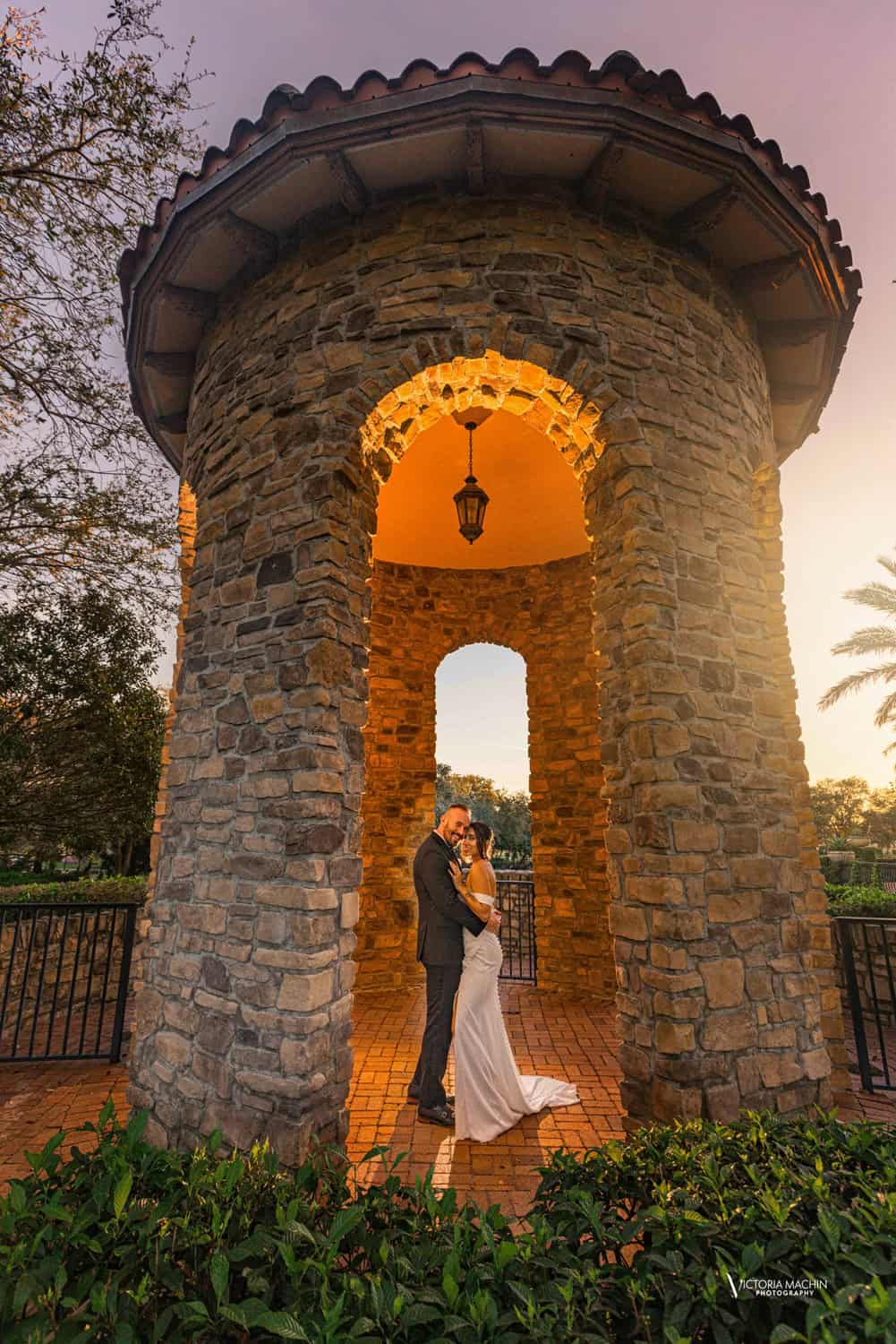 Bride and groom embracing beneath a stone tower lit by a hanging lantern at golden hour, Parkland Golf Club