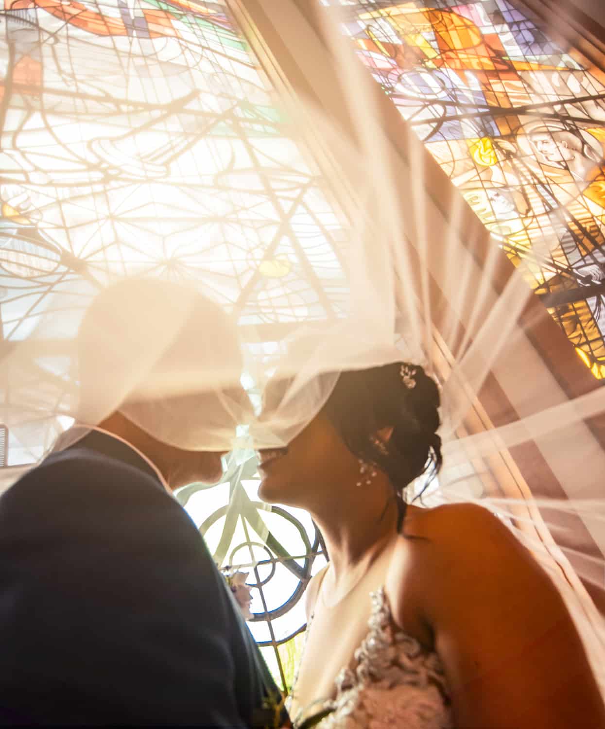 Bride and groom about to kiss beneath her veil, stained glass glowing behind them