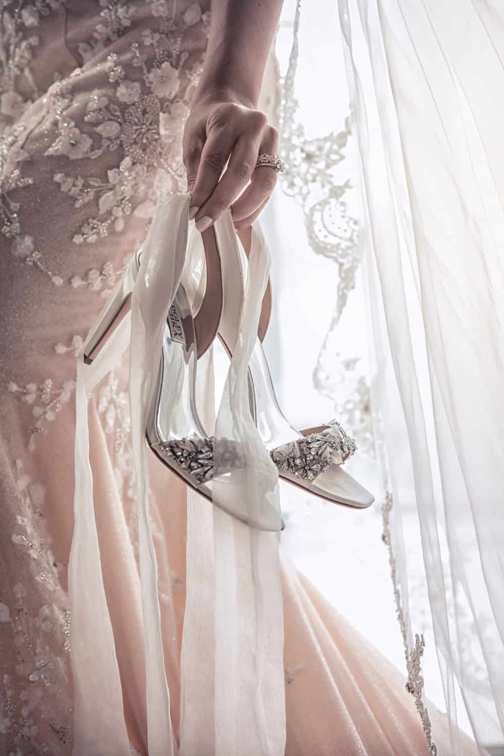 Bride holding her embellished Badgley Mischka heels beside a lace-edged veil in window light