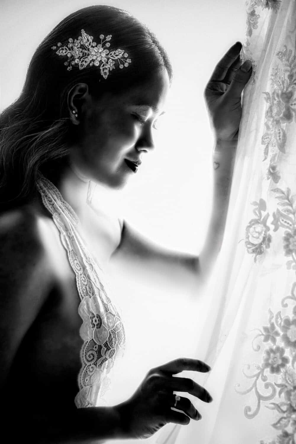 Bride in profile touching a lace curtain in soft window light, in black and white