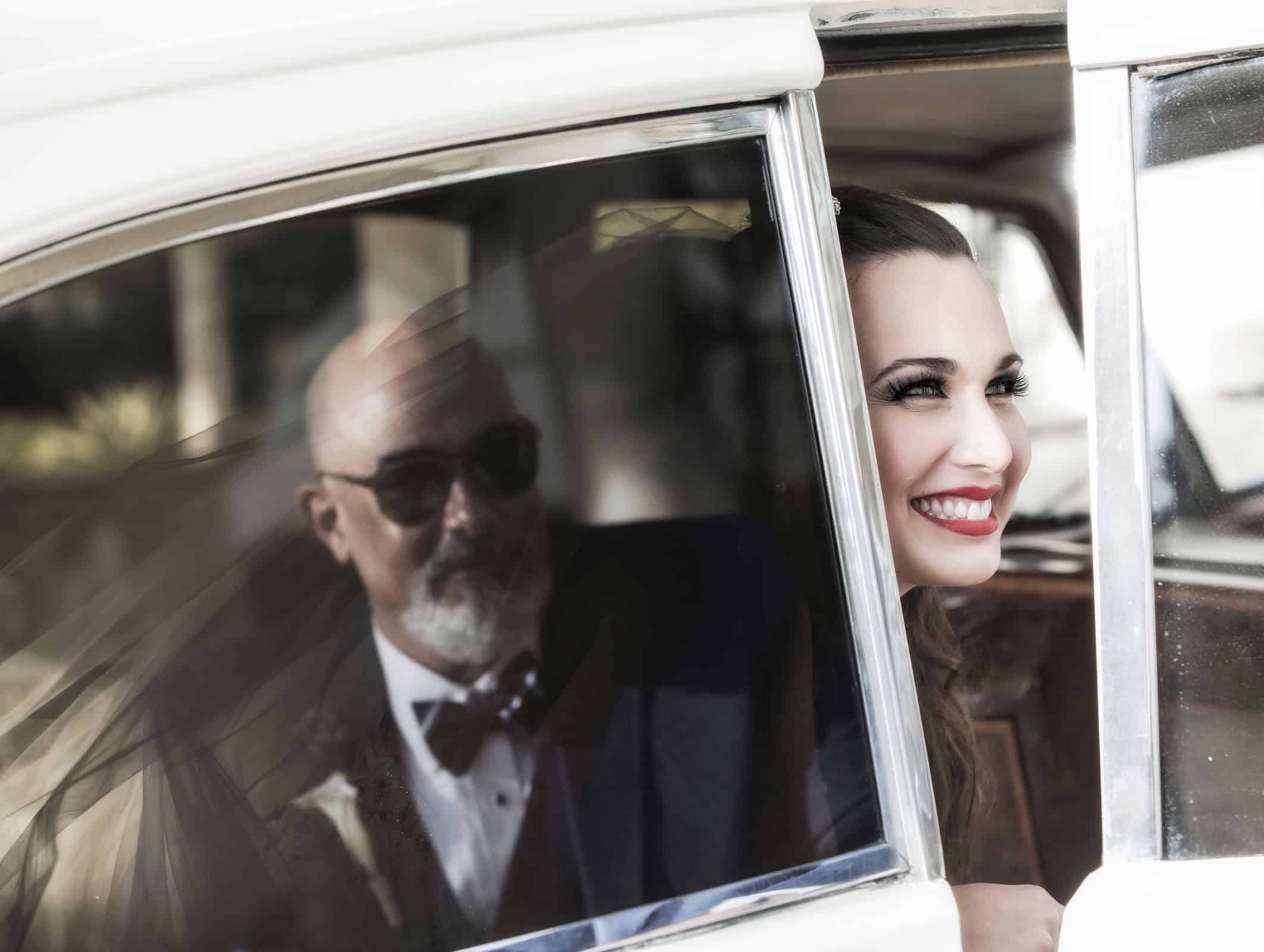 Bride smiling from the window of a vintage white car, her father seated beside her