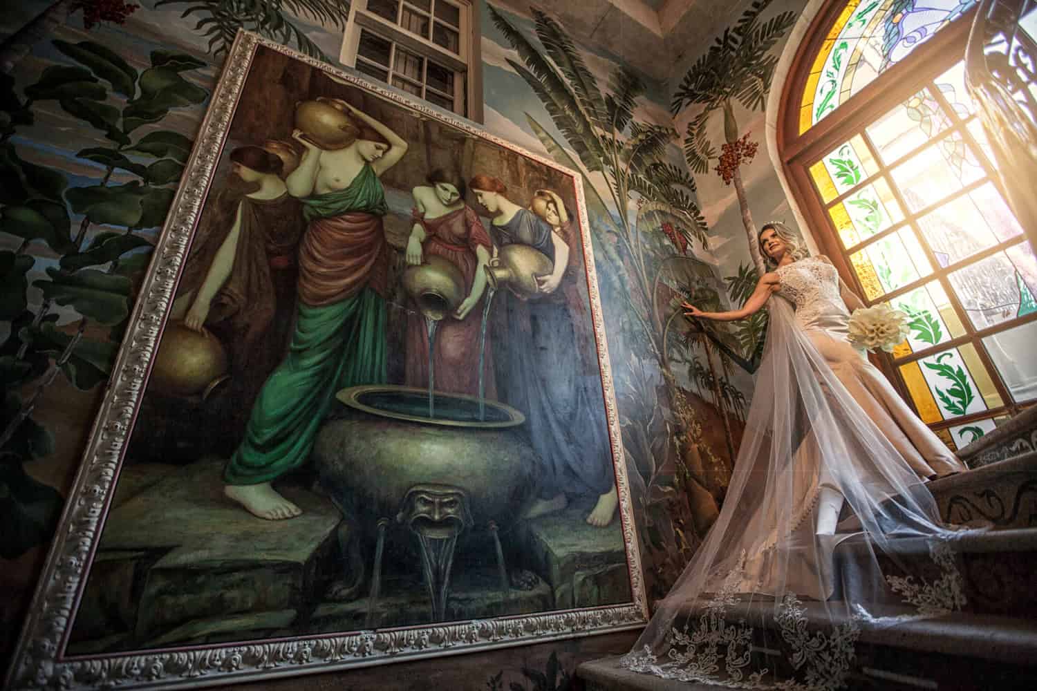 Bride on a grand staircase beside a classical painting and tropical mural, stained-glass window glowing above