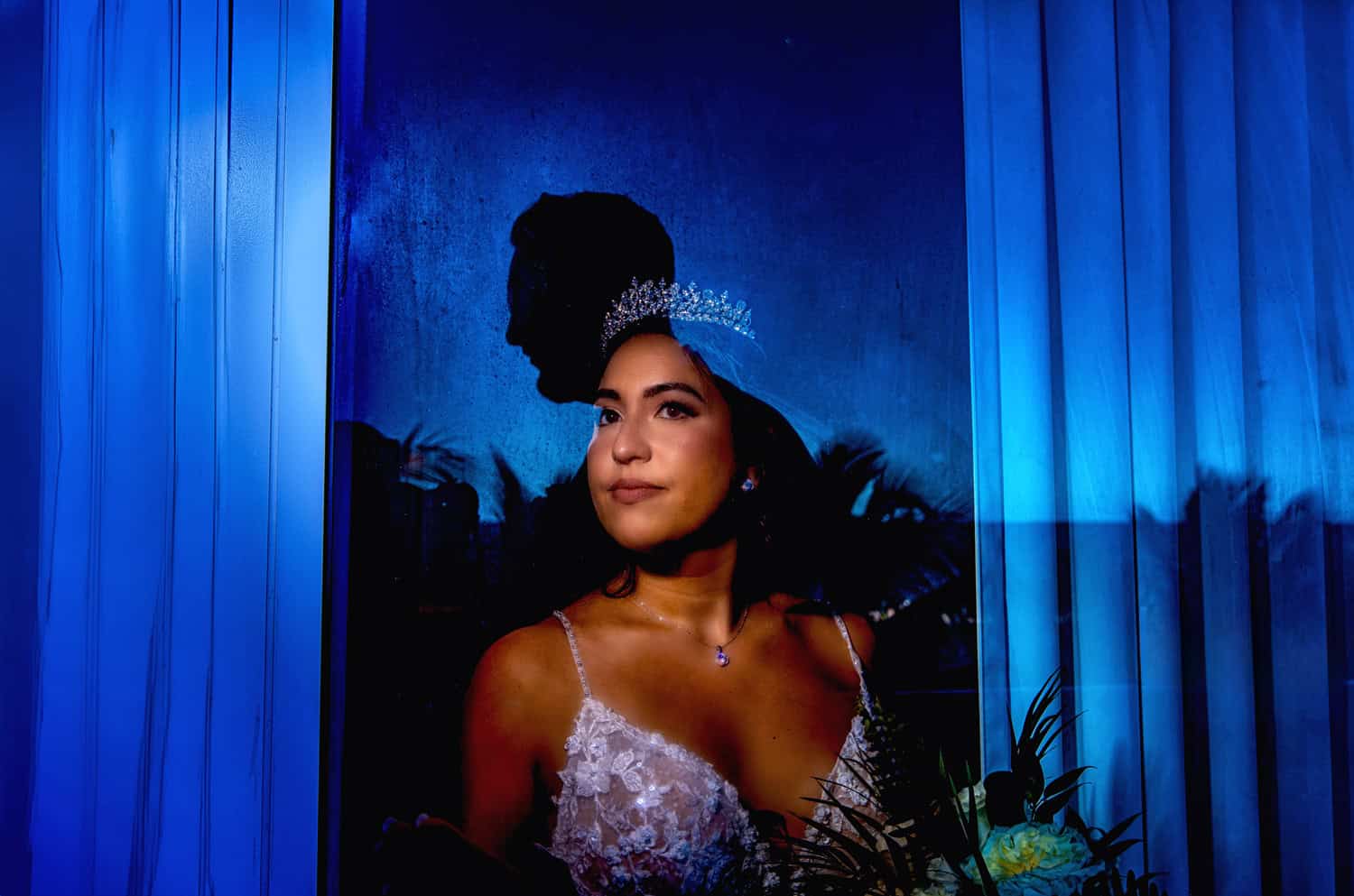 Bride in a crystal tiara lit through a window in blue light, her groom’s silhouette in shadow behind her