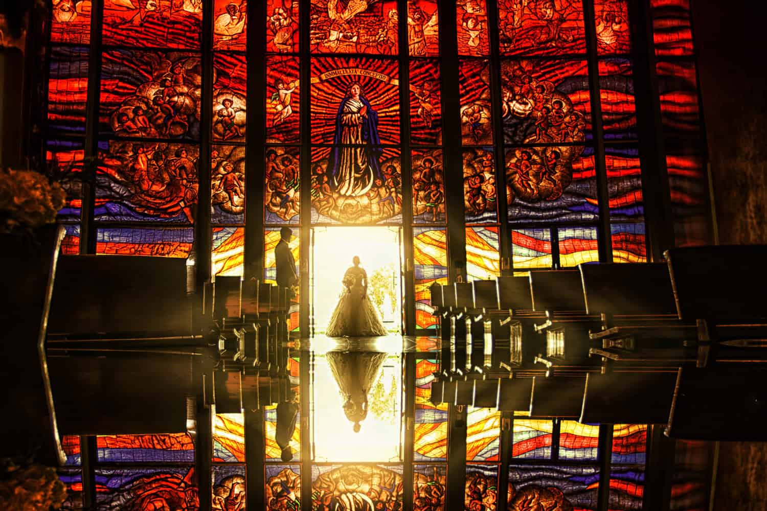Bride and groom silhouetted in a chapel doorway beneath a vast stained-glass window, mirrored in the polished floor