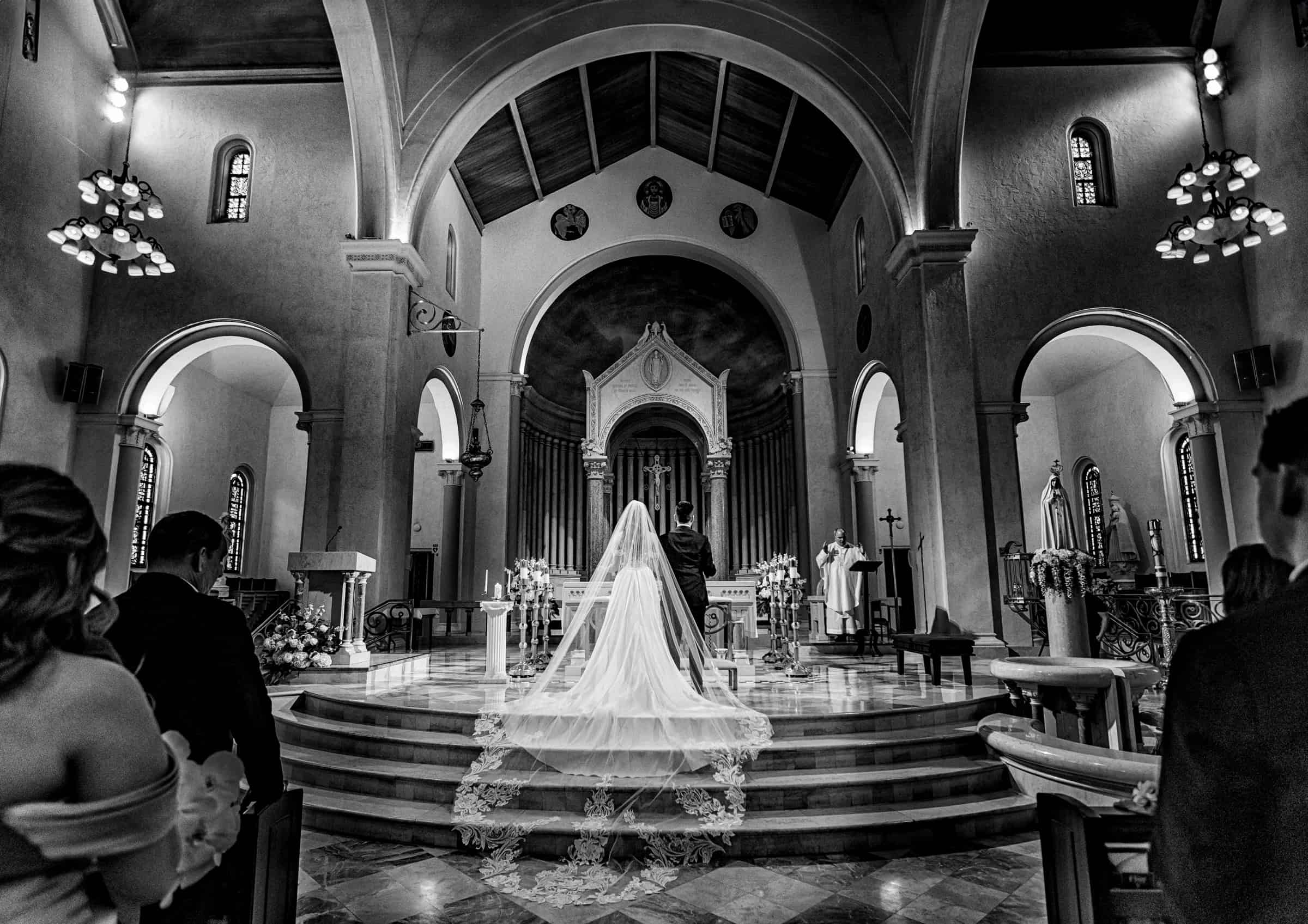 Bride and groom at the altar of a Catholic church, her cathedral veil spilling down the steps, black and white