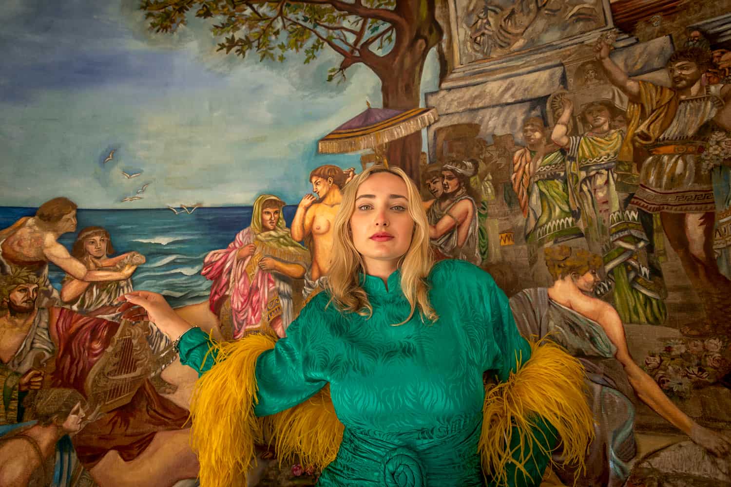 Editorial portrait of a woman in an emerald gown with feathered sleeves before a painted classical mural