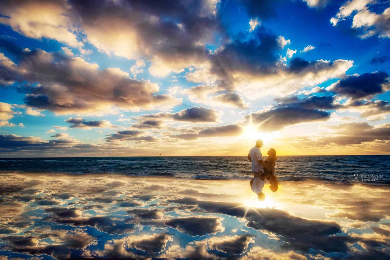 Silhouetted couple at the shoreline beneath a dramatic sunrise sky mirrored in the sand