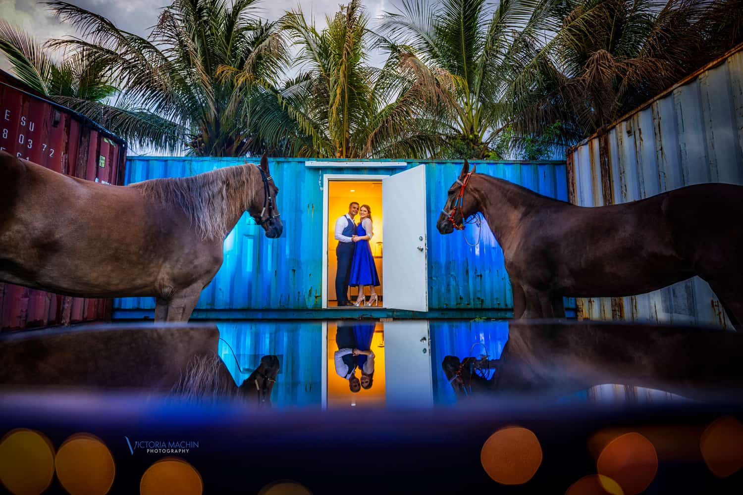 Couple in a glowing doorway between two horses and shipping containers, reflected below