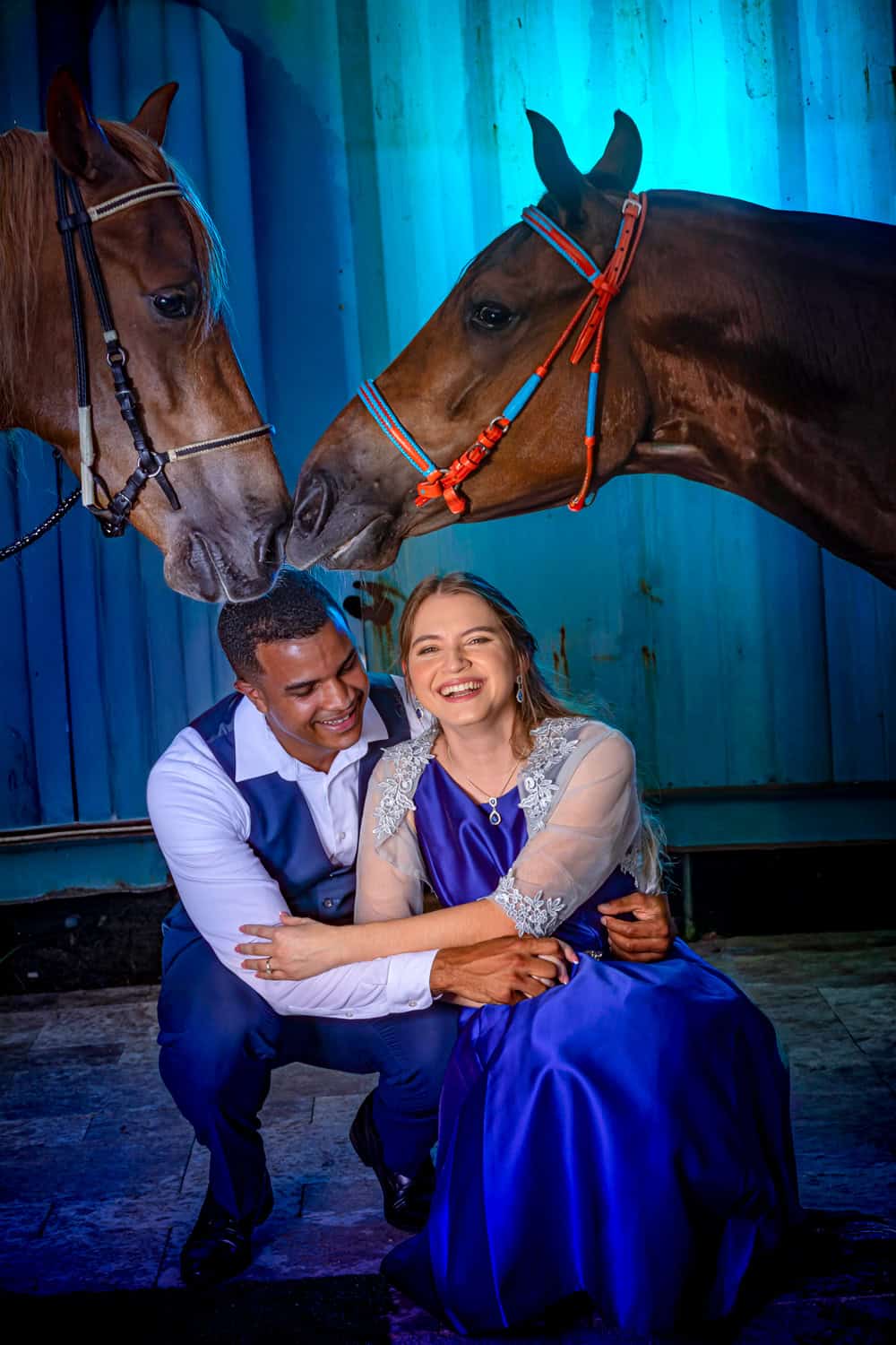 Laughing couple crouched beneath two horses touching noses in blue light