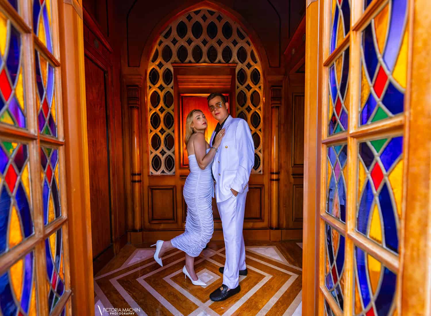 Engaged couple in white framed by stained-glass doors and carved wood arches