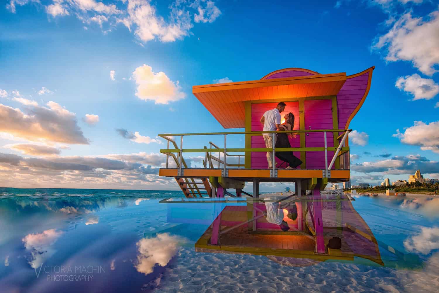 Couple embracing on a pink Miami Beach lifeguard tower at sunrise, reflected in wet sand