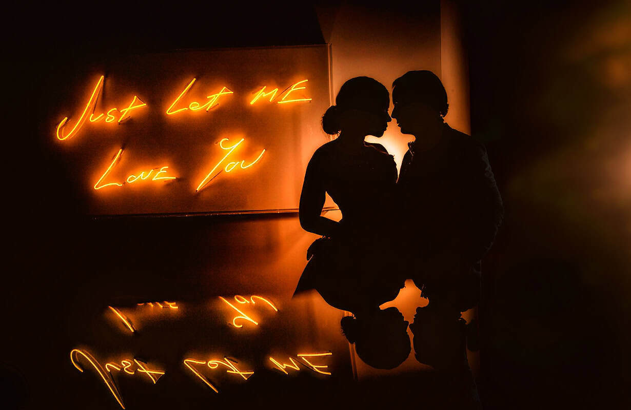 Silhouetted engaged couple nose to nose beside a neon sign reading Just Let Me Love You