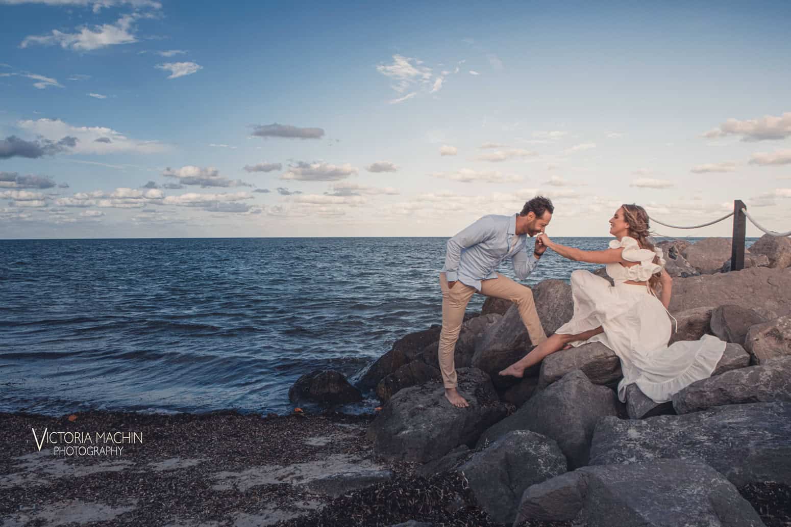 He kisses her hand on the ocean rocks under a blue sky