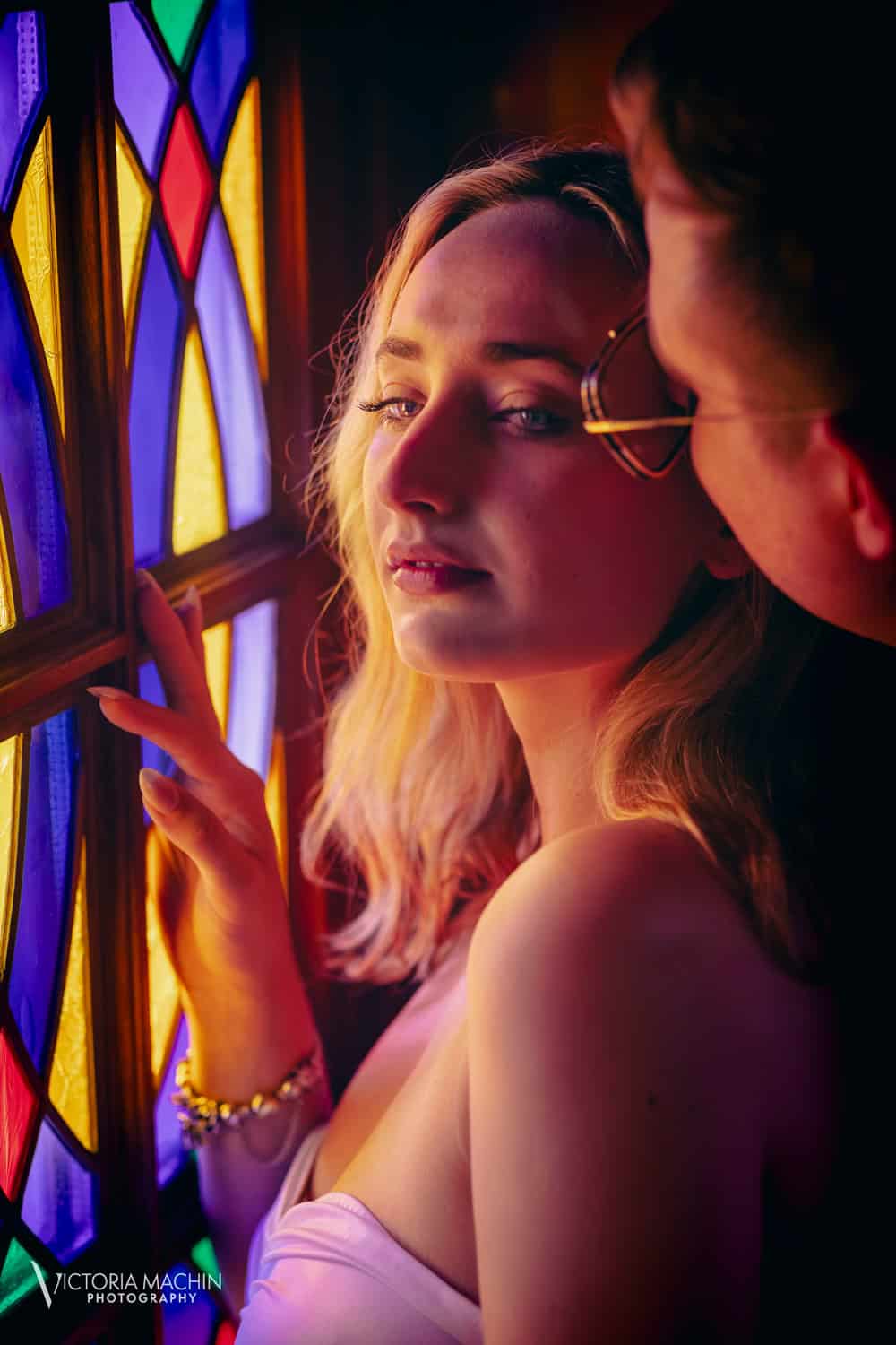 Bride-to-be bathed in stained-glass light as her fiancé holds her from behind