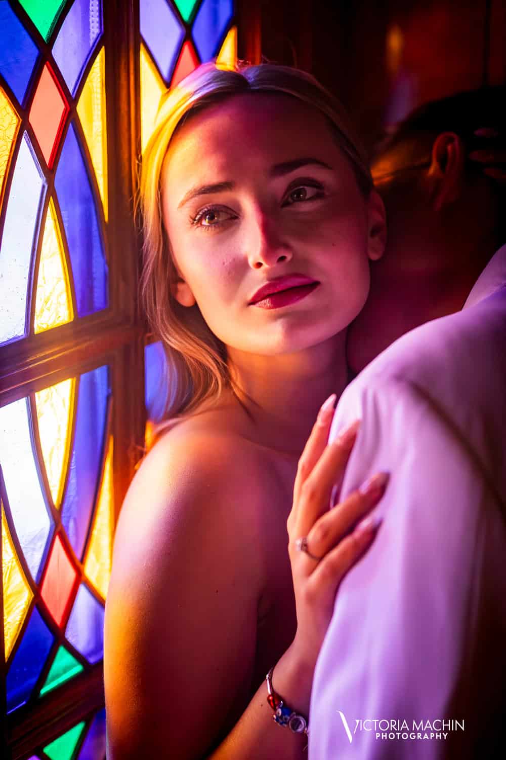 Bride-to-be resting her ringed hand on her fiancé’s shoulder beside a stained-glass window