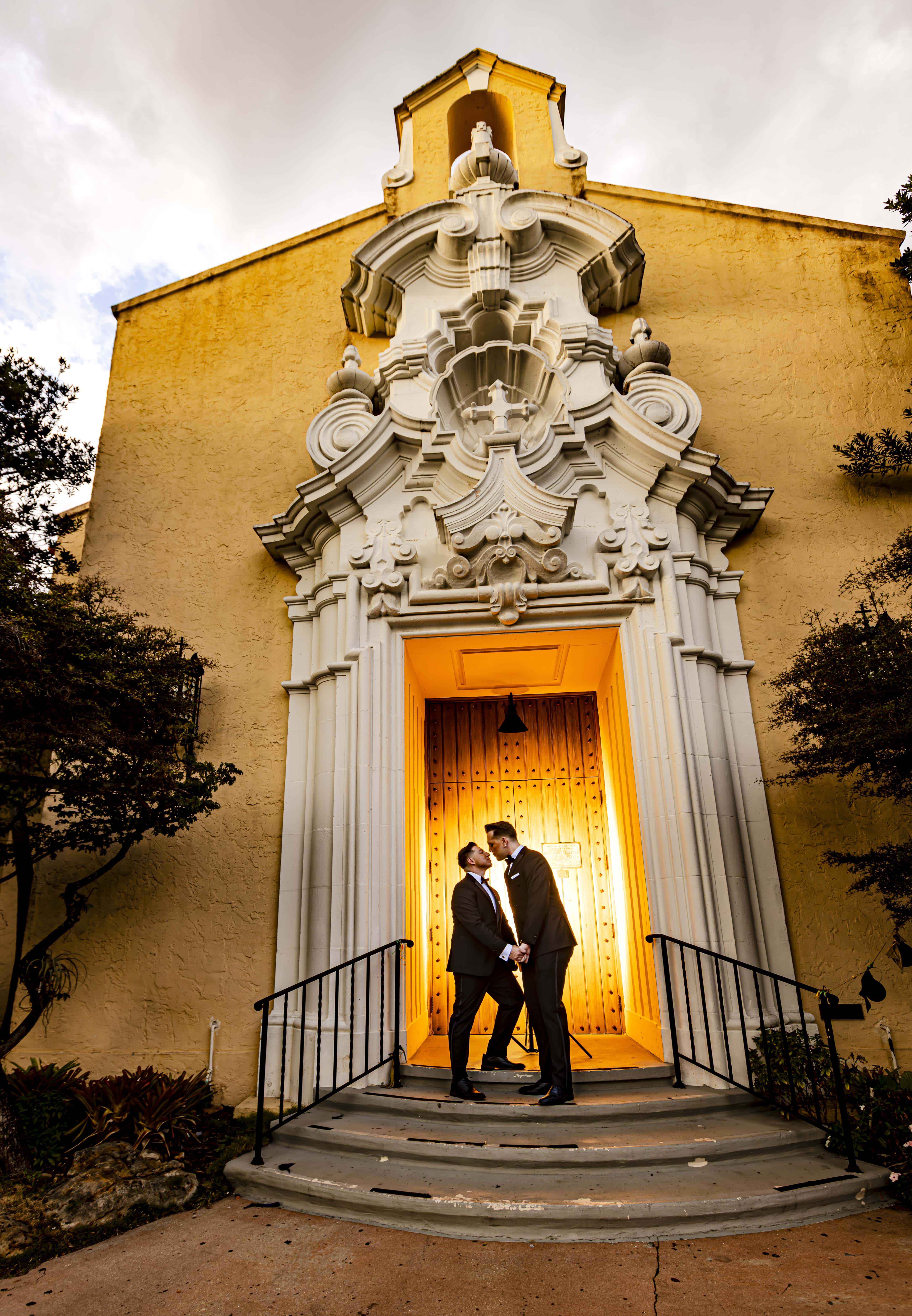 Two grooms about to kiss in the golden-lit doorway of a Spanish baroque church