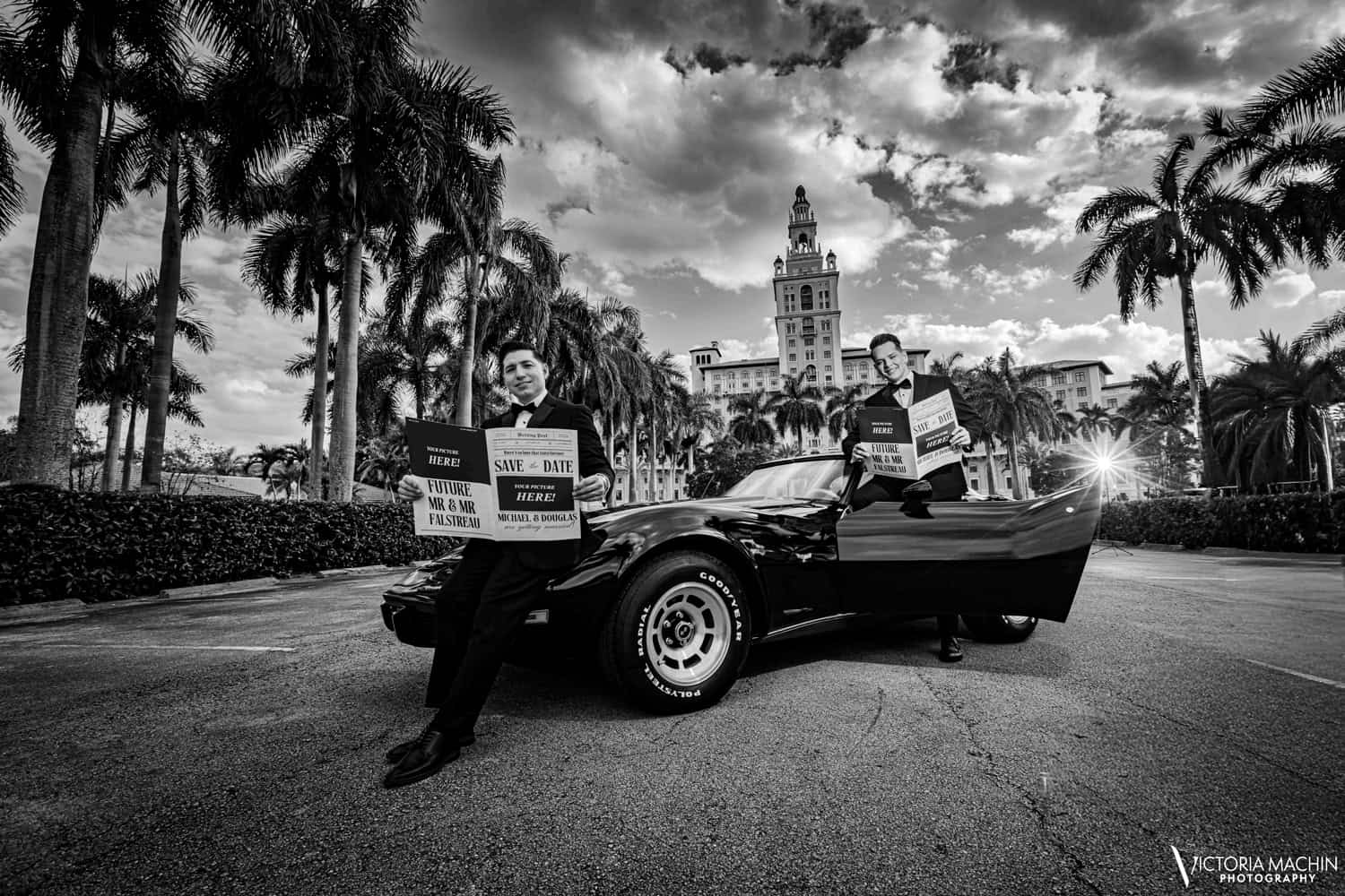 Two grooms in tuxedos with save-the-date newspapers beside a vintage Corvette at The Biltmore, in black and white
