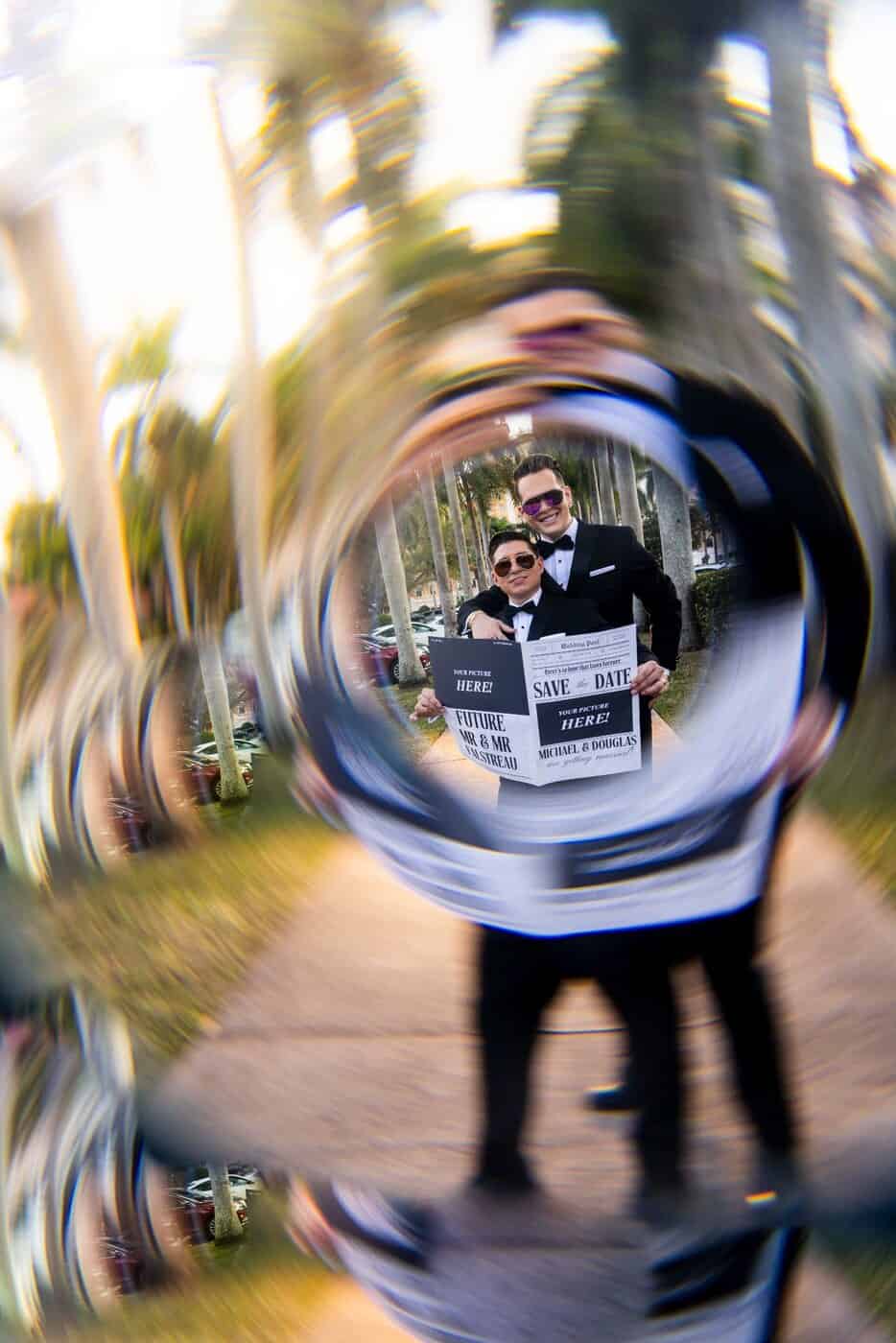 Two grooms holding save-the-date newspapers, photographed through a glass reflection
