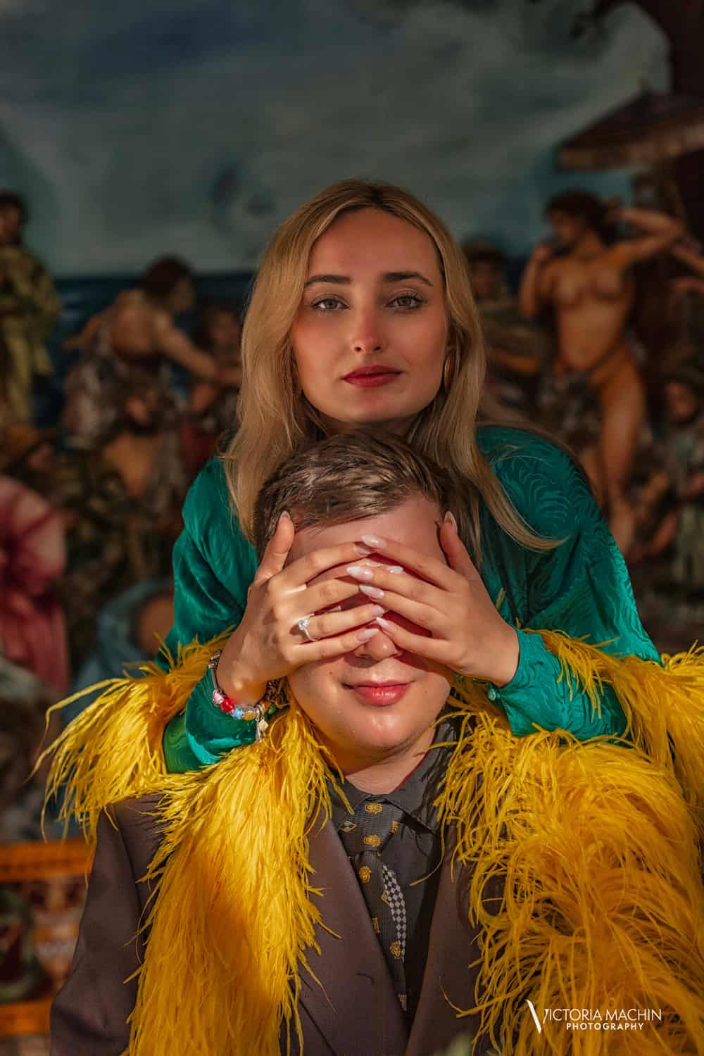 Bride-to-be covering her fiancé’s eyes, ring showing, before a painted mural at the Versace Mansion, he in a yellow feather boa