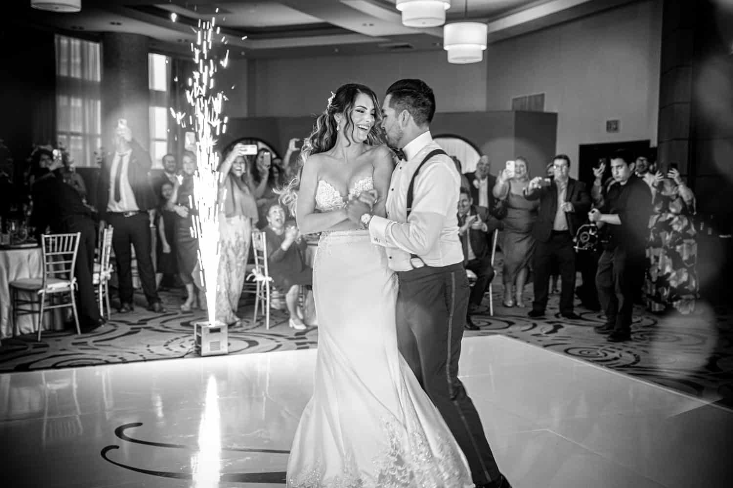Bride and groom laughing during their first dance as sparklers light the ballroom, in black and white