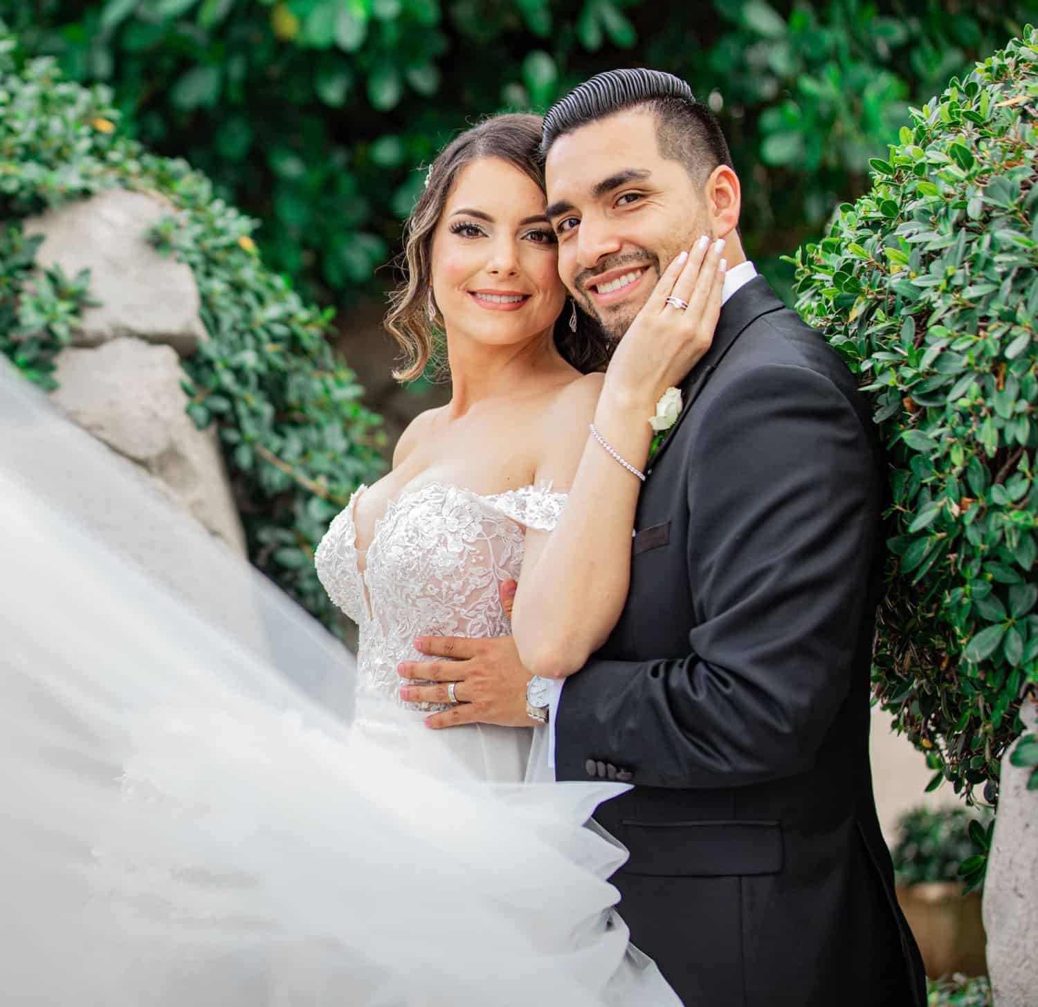 Bride and groom cheek to cheek among lush greenery, her veil sweeping into the frame