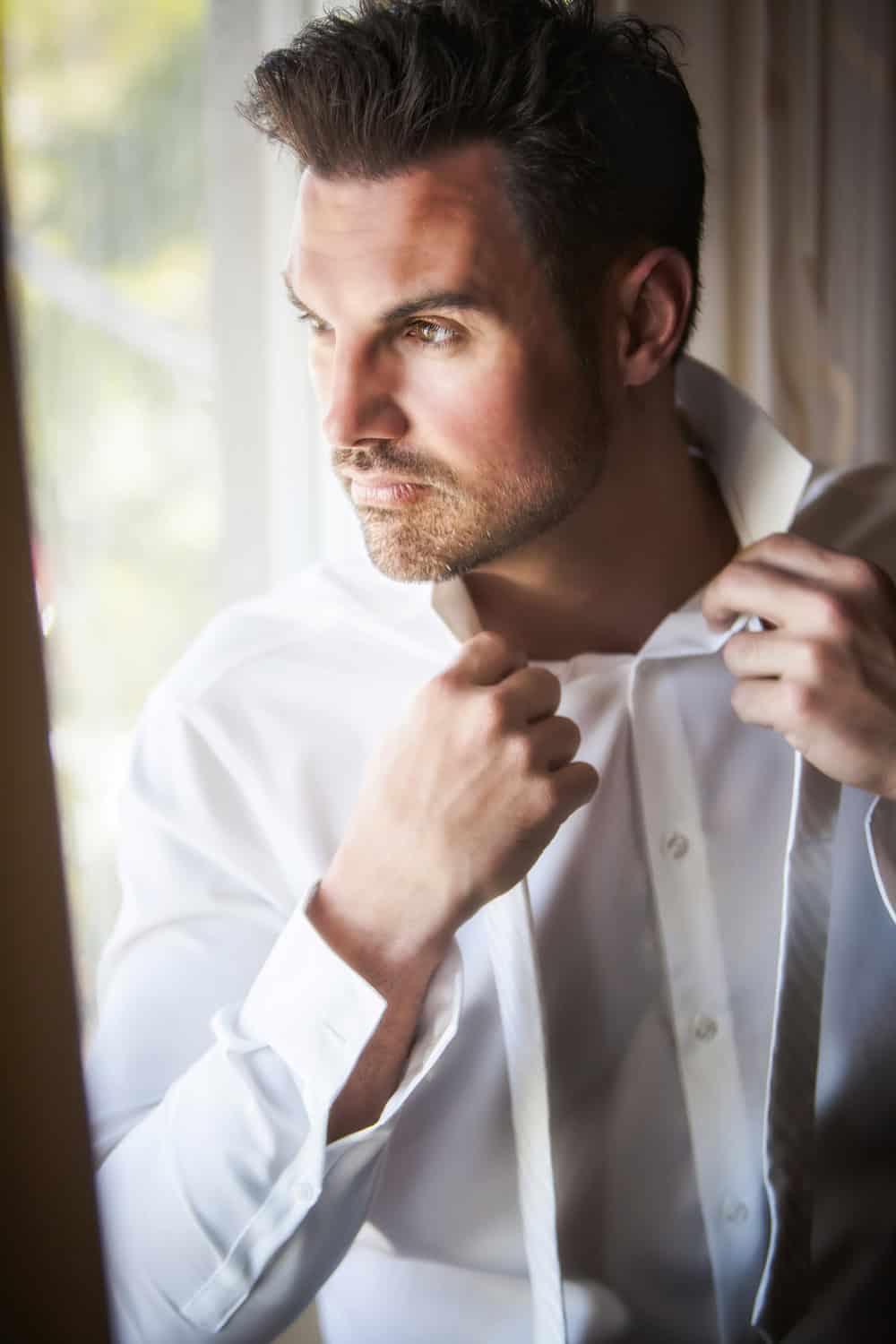 Groom adjusting his collar in window light while getting ready