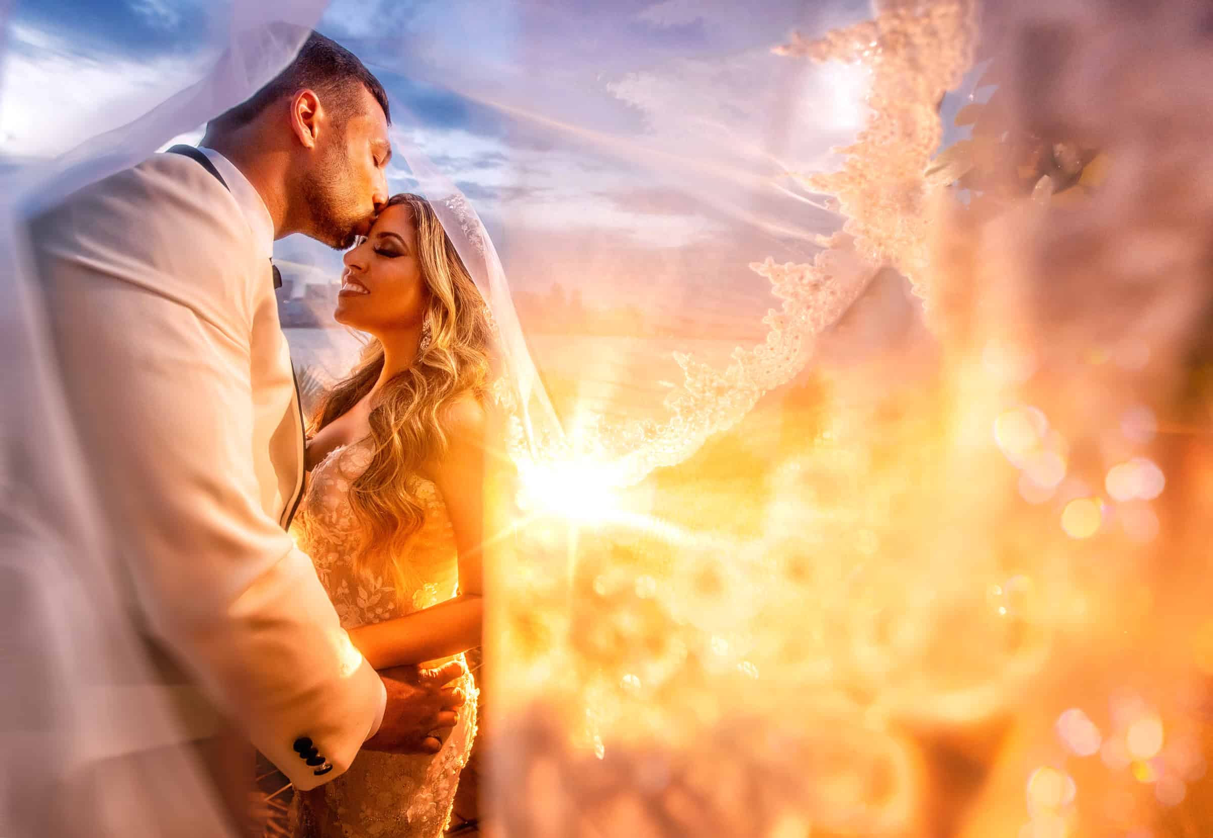 Groom kissing the bride’s forehead beneath her veil as the sun sets over the water