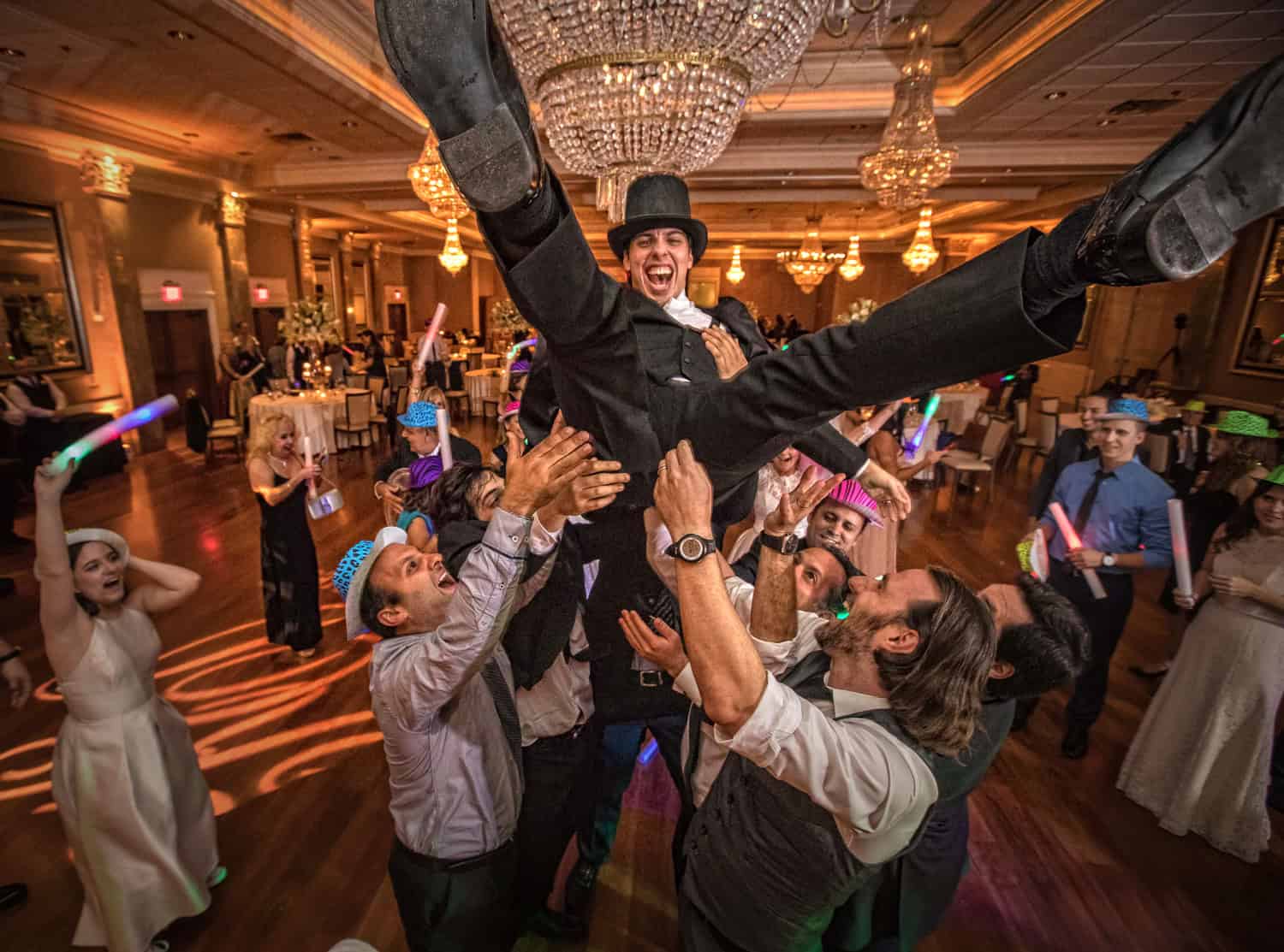Groom in a top hat lifted by his friends beneath ballroom chandeliers