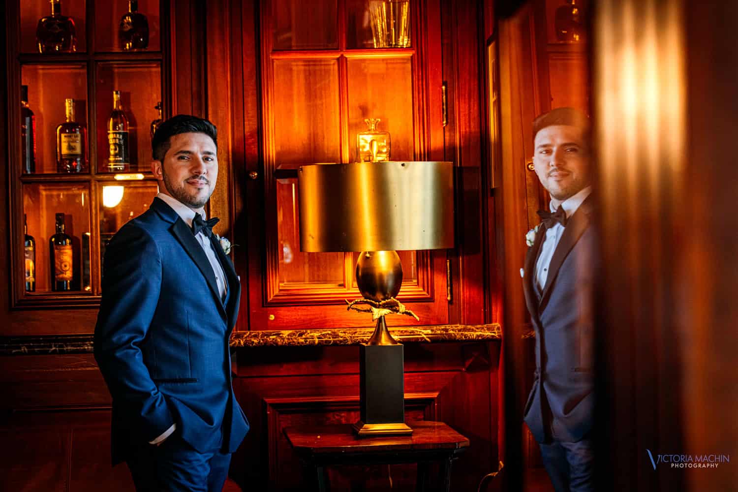 Groom in a navy tuxedo in a wood-paneled whisky bar, reflected in the glass