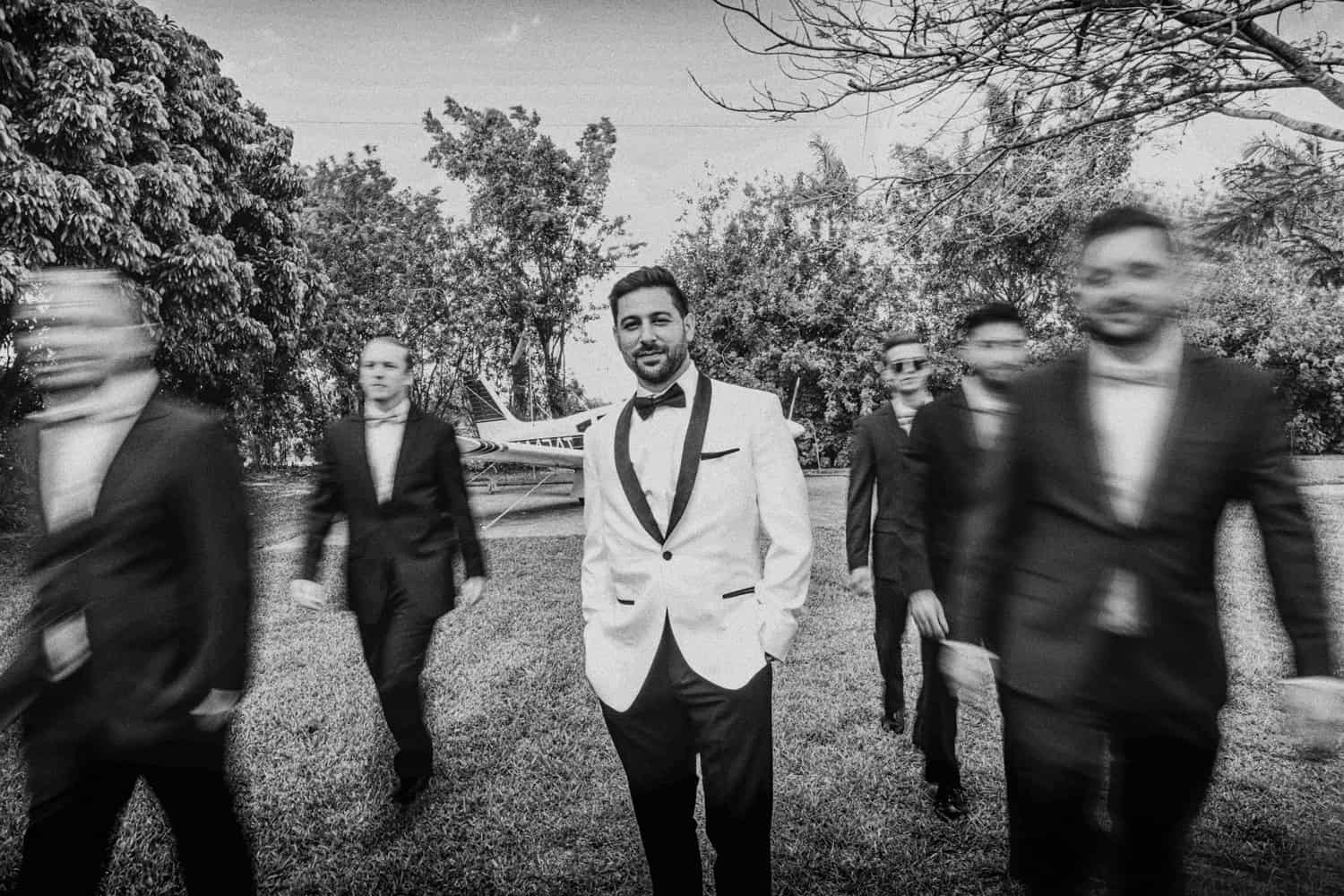 Groom in a white dinner jacket standing still as his groomsmen walk past in motion blur, black and white