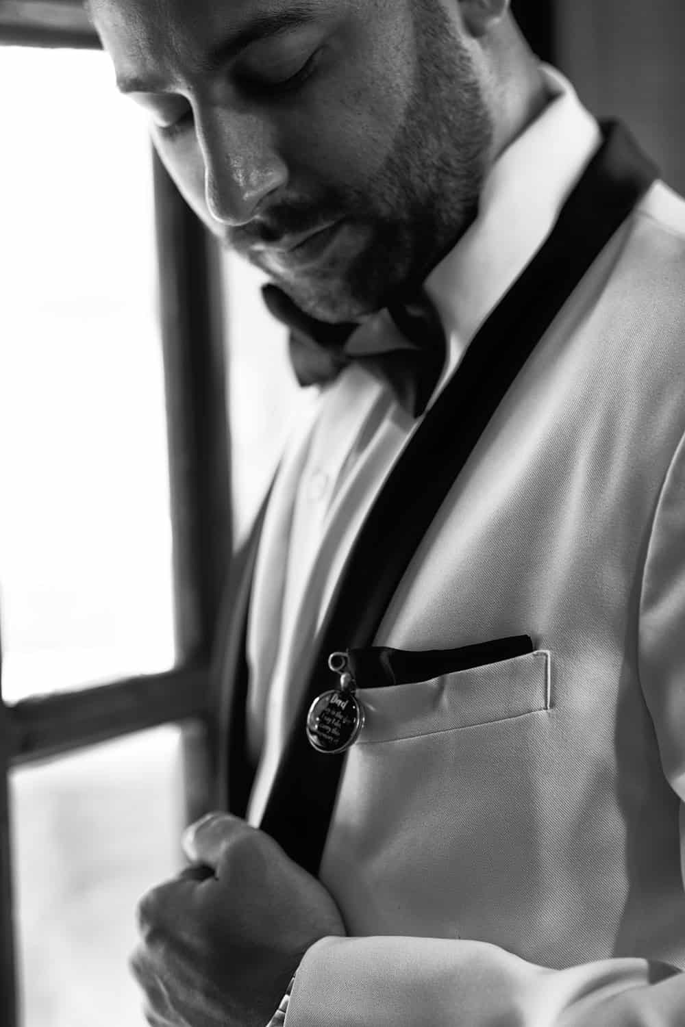 Groom in a white tuxedo looking down at a memorial charm pinned to his pocket square, black and white