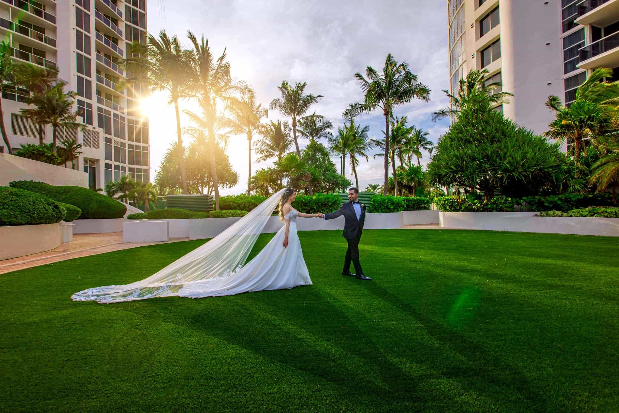 Bride and groom holding hands on a sunlit lawn framed by palm trees in Miami