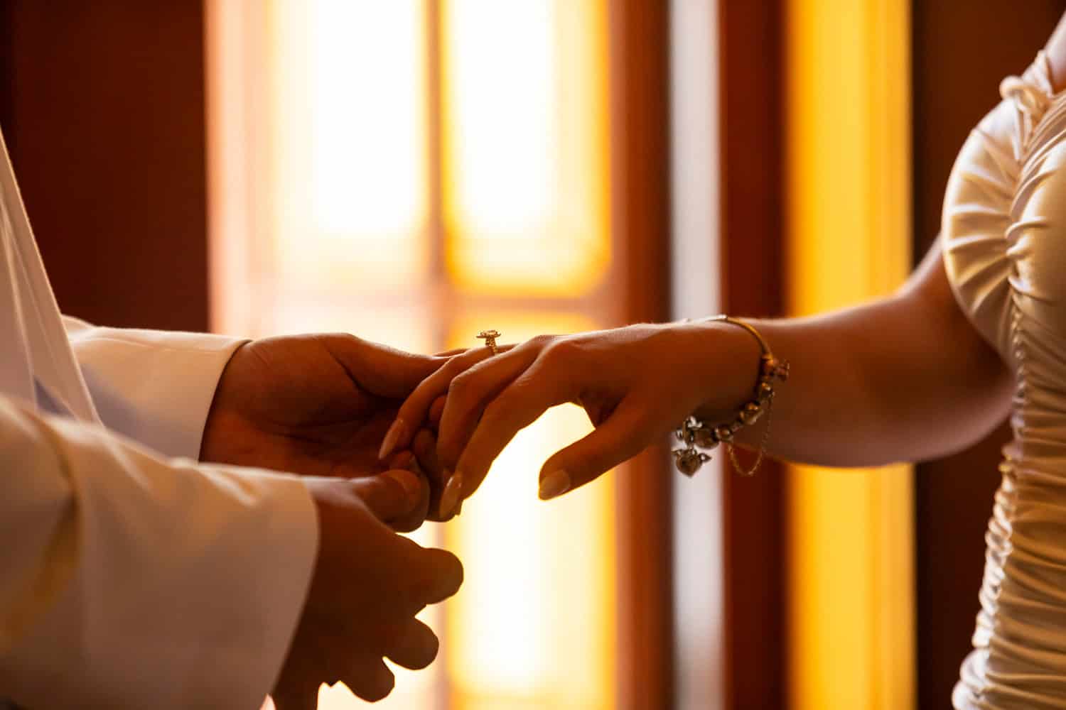 Close-up of an engagement ring as he holds her hand before a glowing window