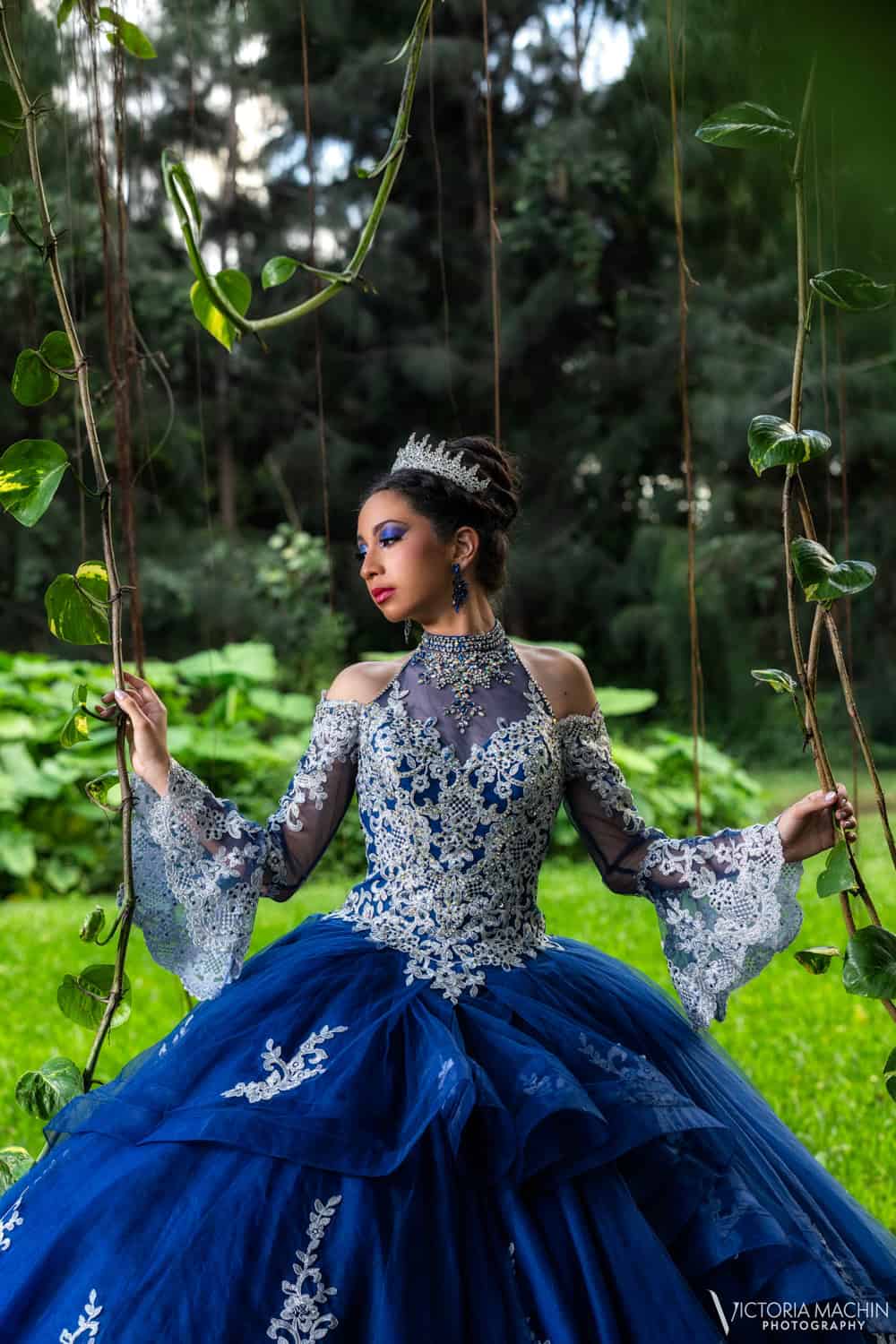 Quinceañera in a royal blue lace gown and crown on a vine swing in a tropical garden