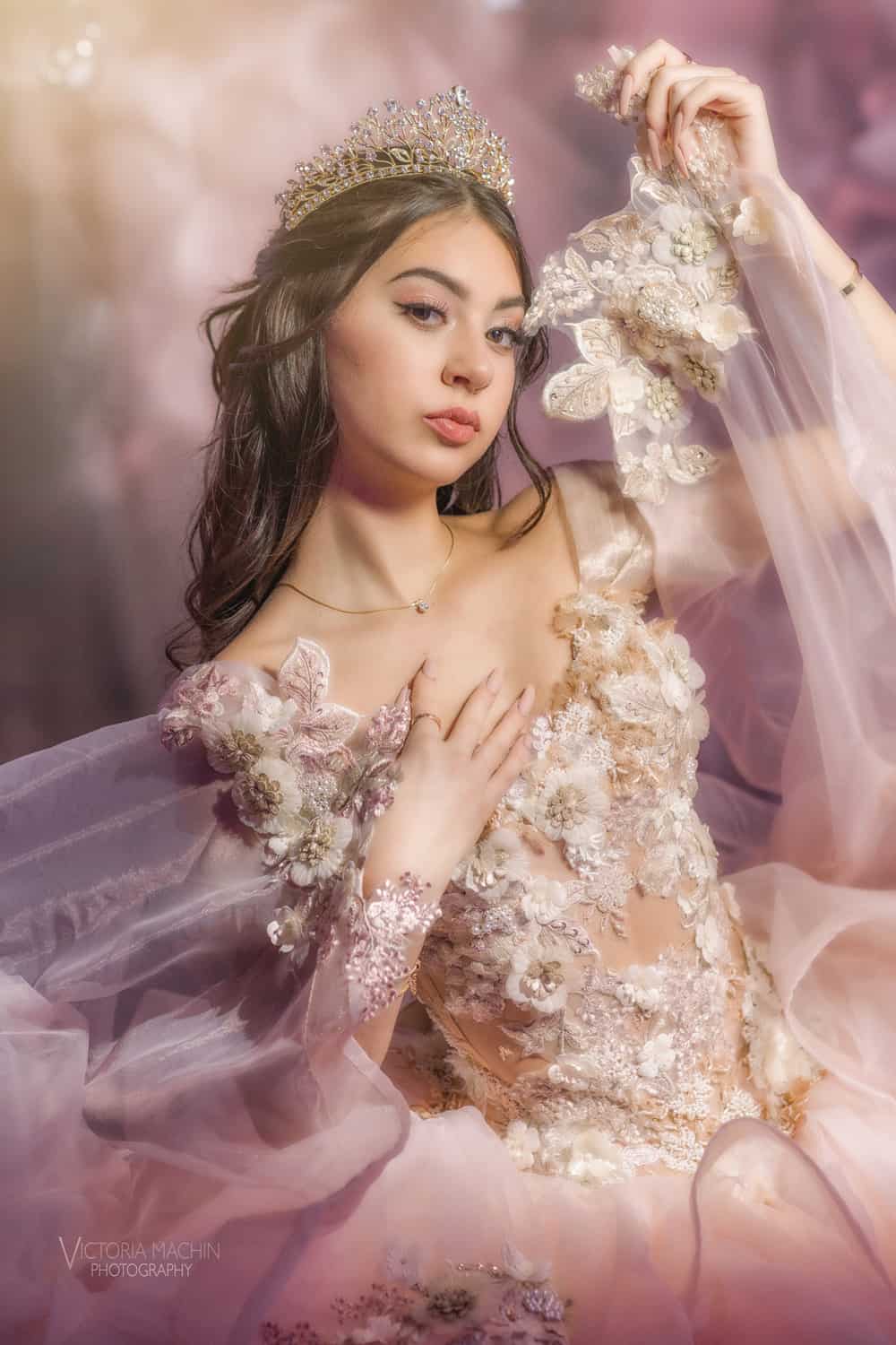 Quinceañera in a tiara and floral-embroidered gown, editorial studio portrait in soft pink light