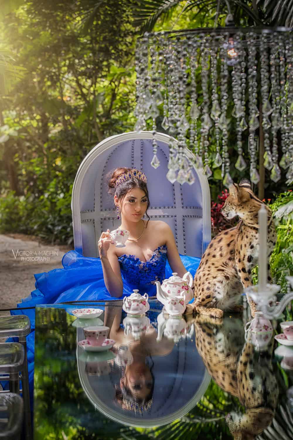 Quinceañera in a royal blue gown at a garden tea party beneath a crystal chandelier, a spotted serval cat beside her
