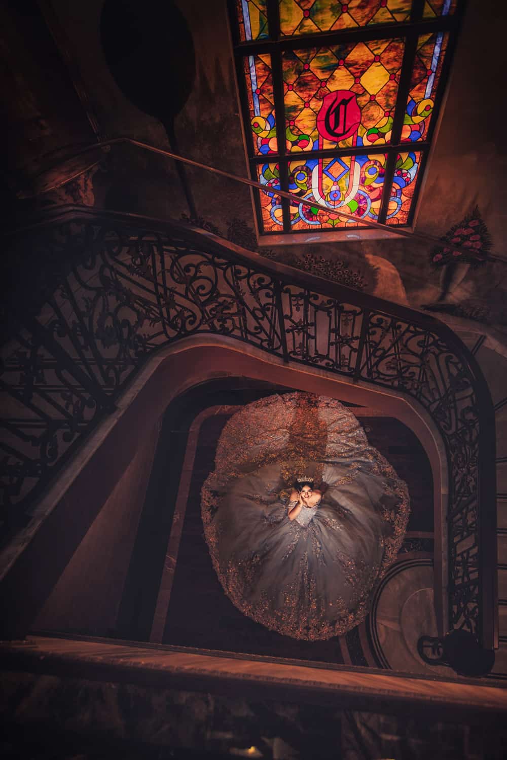 Quinceañera in a full ball gown photographed from above at the foot of a grand spiral staircase beneath a stained-glass skylight