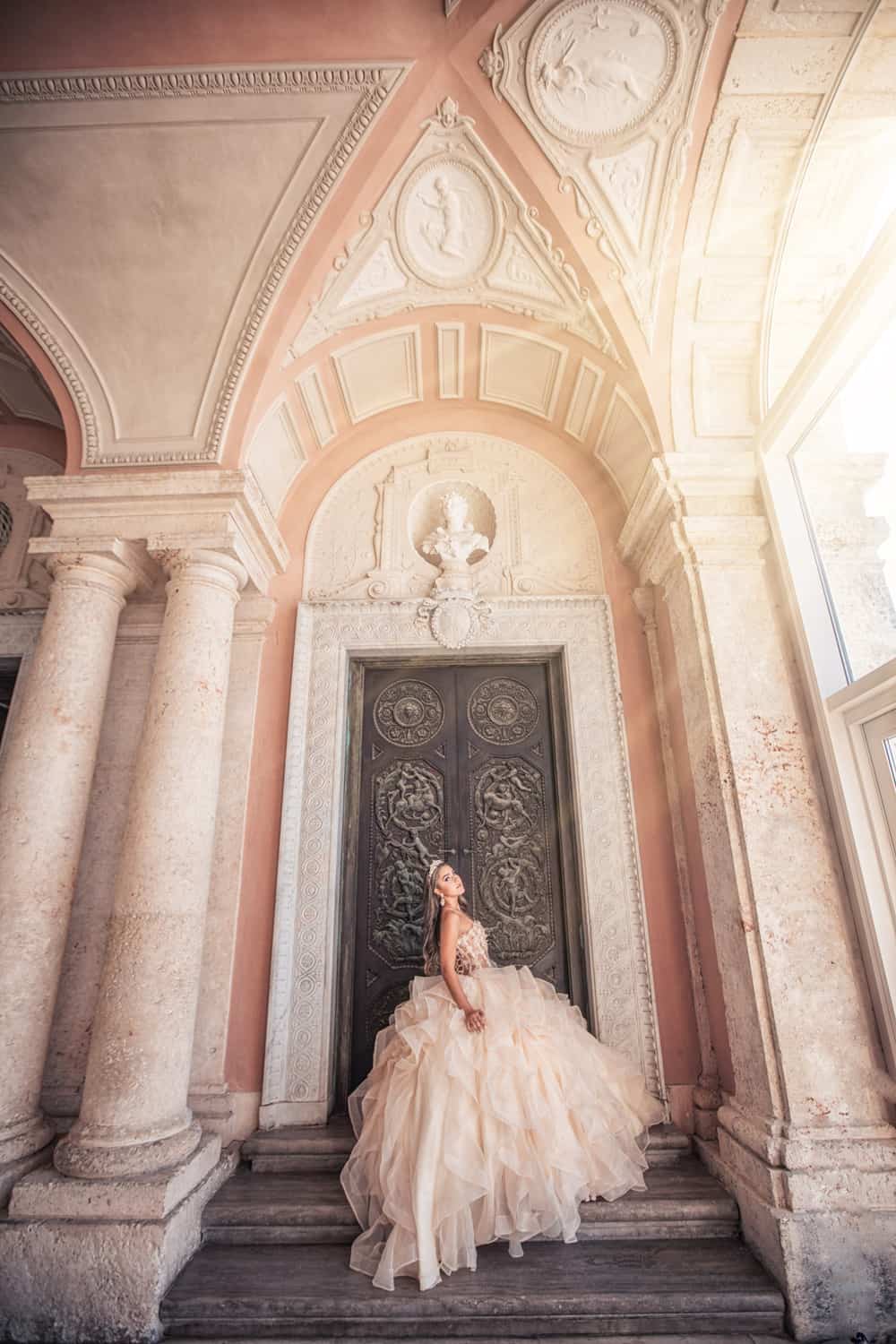 Quinceañera in a blush ruffled gown on the steps of an ornate Mediterranean archway